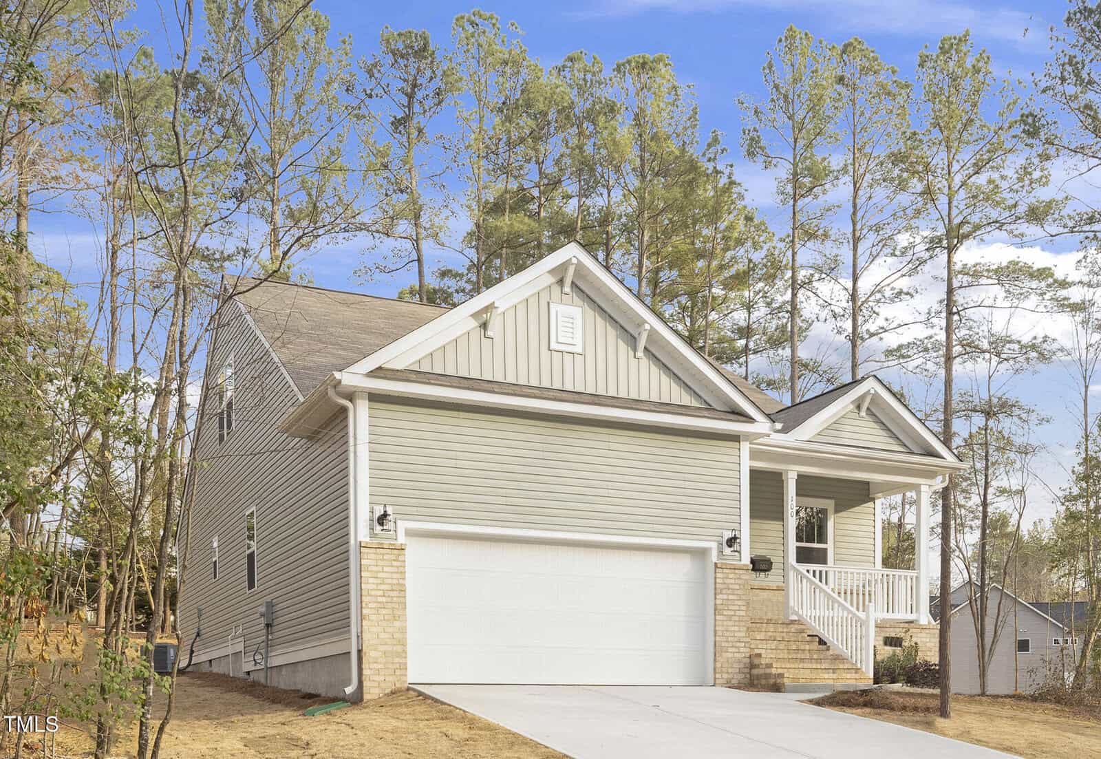 A modern light gray house with white trim, a covered porch, brick accents, and a double garage sits on a sloped lot with a concrete driveway, surrounded by tall pine trees under a blue sky.