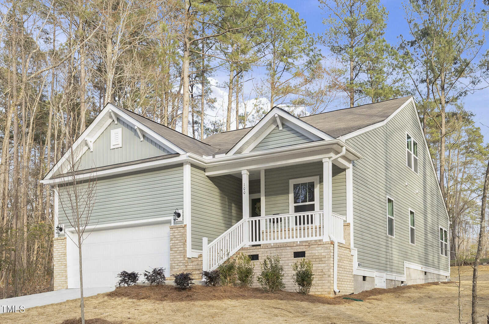 A single-story house with green siding, white trim, and a small front porch. The home has a two-car garage and is surrounded by tall trees, with dry grass and minimal landscaping in the front yard.