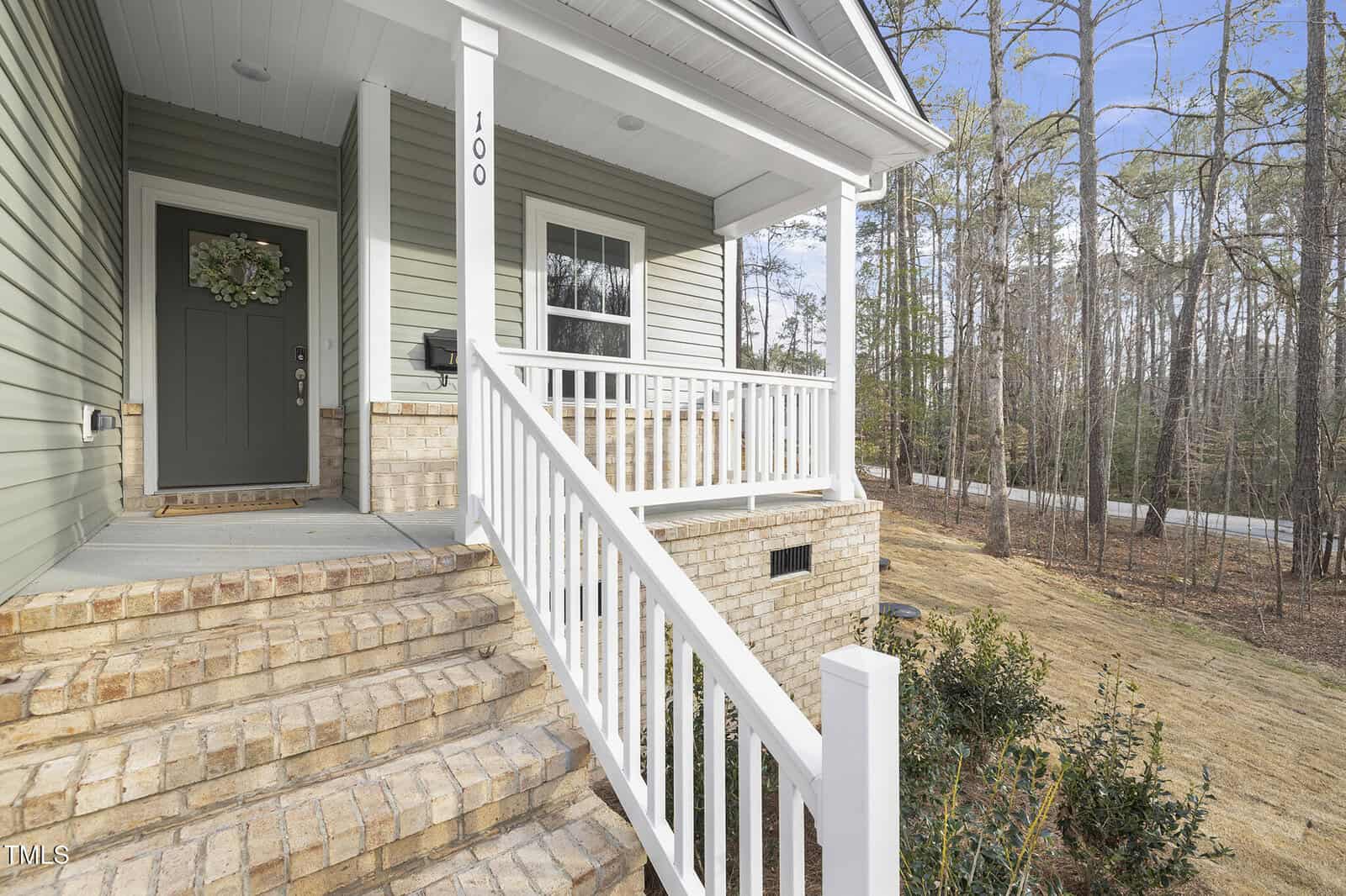 Front porch of a house with light green siding, white trim, a covered entrance, steps leading up with brick detailing, white railing, a dark green door with a wreath, and a wooded area in the background.