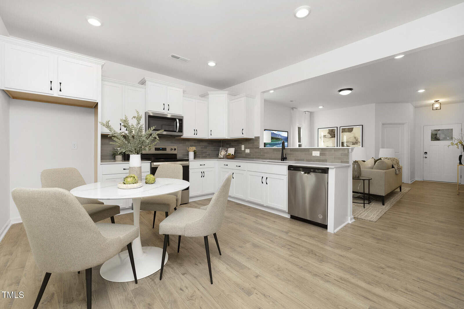 Modern open-concept kitchen and living area with white cabinets, stainless steel appliances, round dining table with four beige chairs, and light wood flooring. The living room features a sofa and framed wall art.