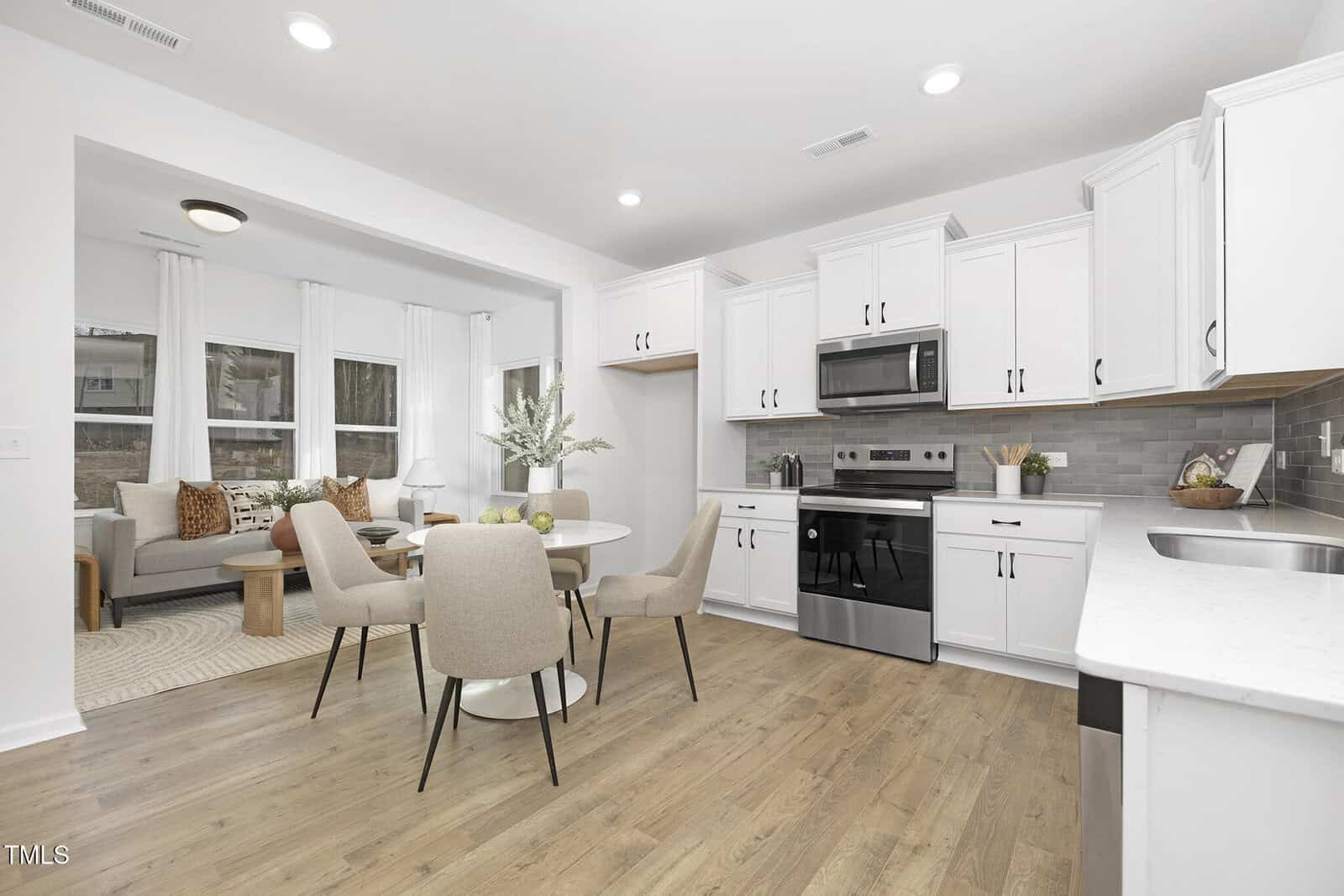 Modern kitchen and dining area with white cabinets, stainless steel appliances, light wood flooring, and a round table with four beige chairs. A living room with a sofa and coffee table is visible in the background.