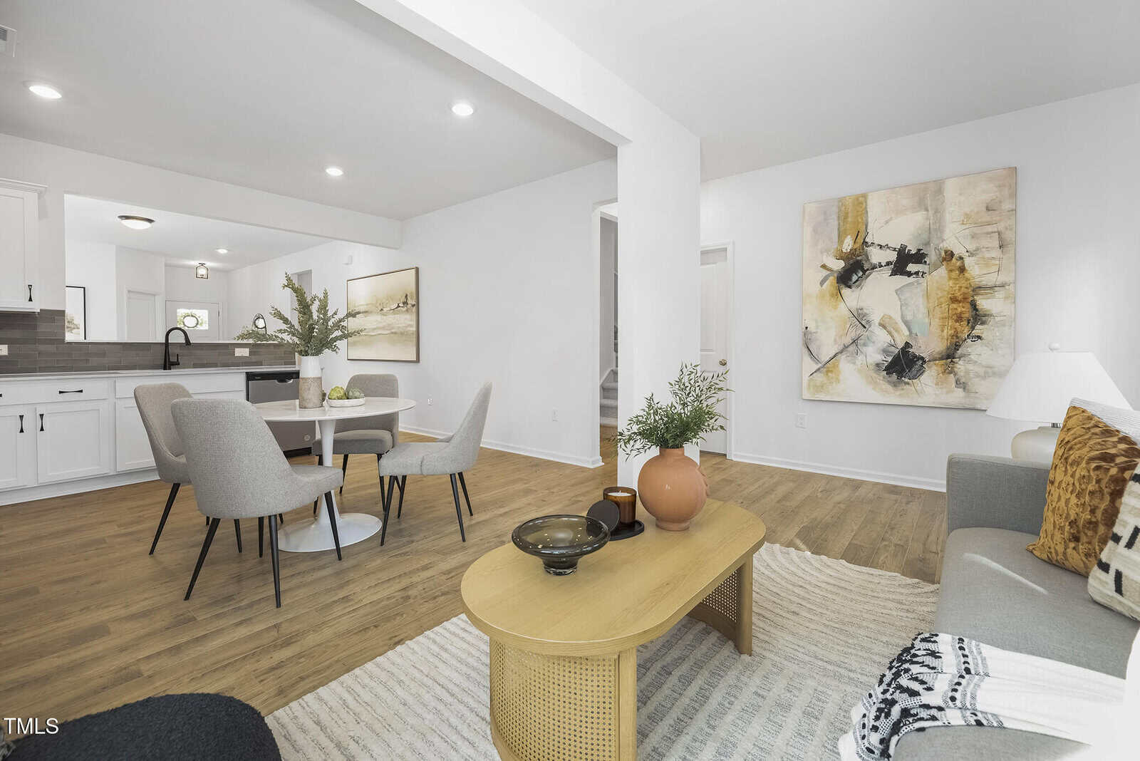 Open-concept living and dining area with wood flooring, white walls, modern furnishings, round dining table, chairs, sofa, coffee table with decor, two abstract paintings, and a kitchen visible in the background.