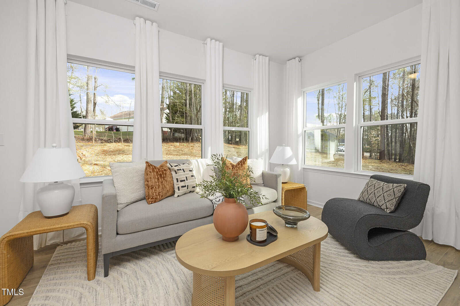 A modern living room with a gray sofa, black lounge chair, wooden coffee table with decor, white lamps, and large windows with white curtains showing a wooded outdoor view.