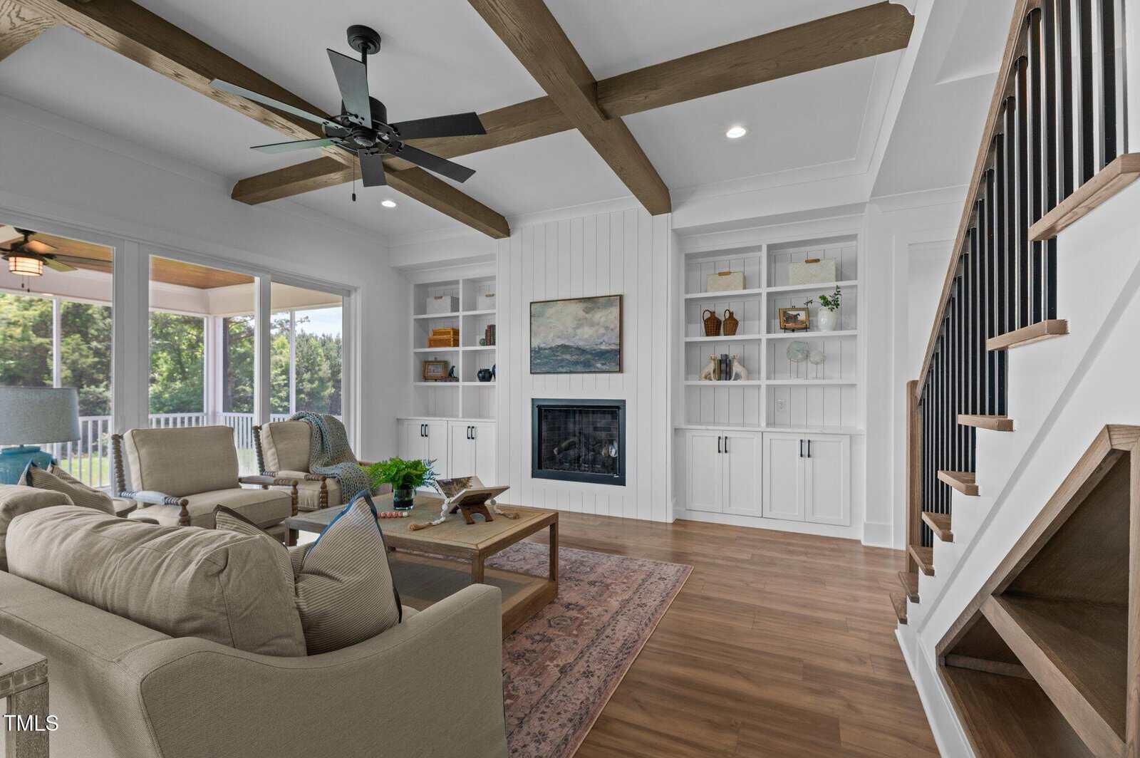 Modern living room with beige sofas, wooden floor, exposed ceiling beams, built-in shelves, a fireplace, and large windows letting in natural light; staircase with black railing on the right.