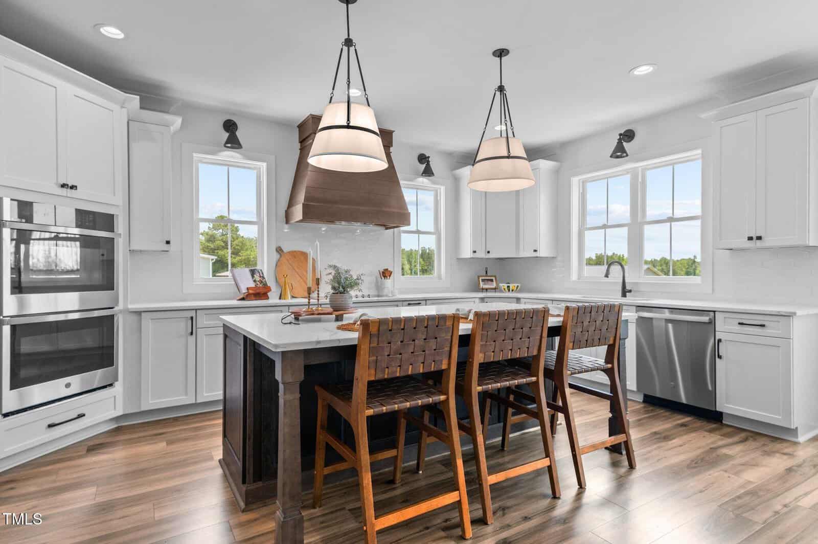 Modern kitchen with white cabinets, stainless steel appliances, a large wooden range hood, island with a dark countertop, and four woven barstools. Two pendant lights hang above the island, and windows let in natural light.