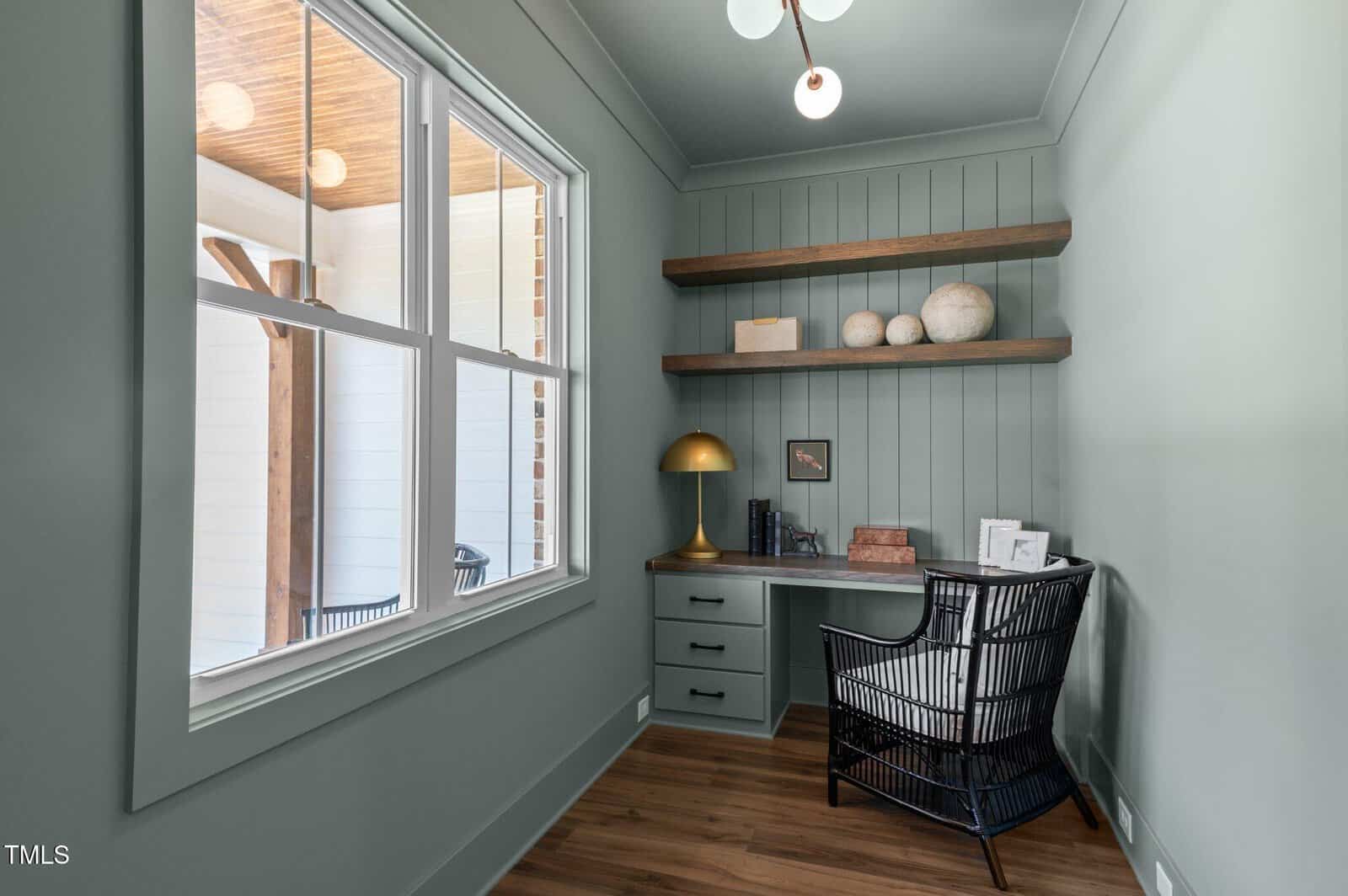 A small home office with sage green walls, a desk with drawers, a black wicker chair, a brass lamp, floating shelves with decor items, wood flooring, and a large window overlooking a porch.