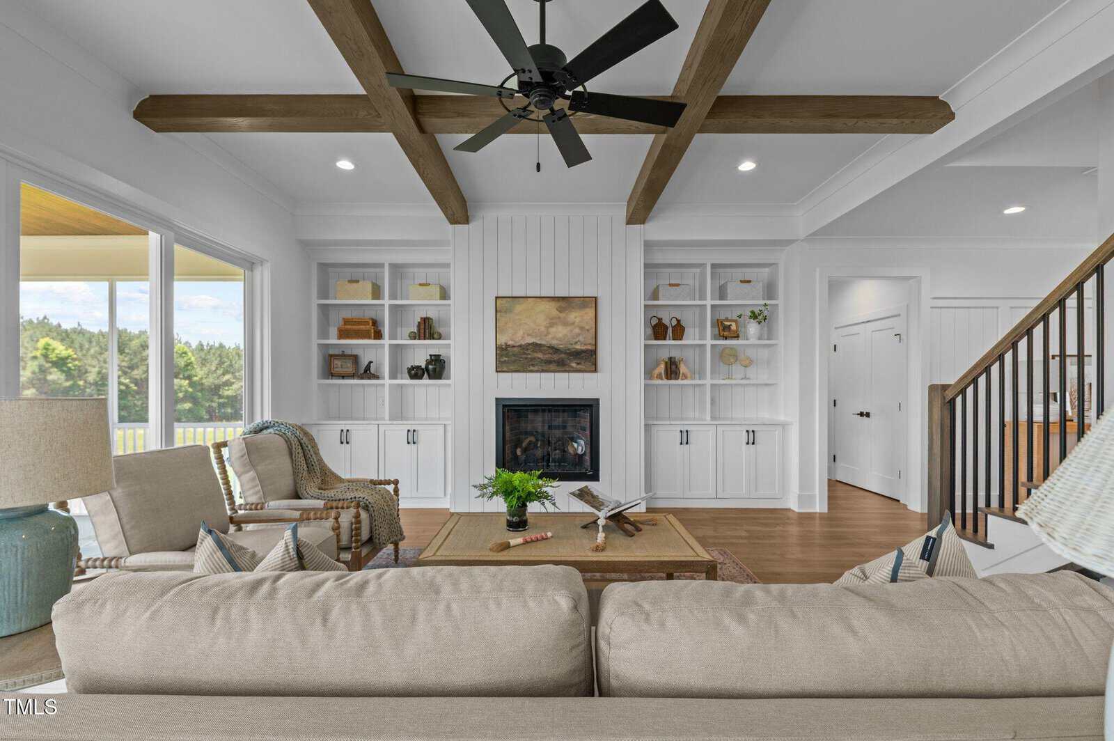 A living room with beige sofas, a coffee table, and built-in shelves around a fireplace. Wooden ceiling beams and a black ceiling fan are overhead. Stairs are visible to the right, and large windows show greenery outside.
