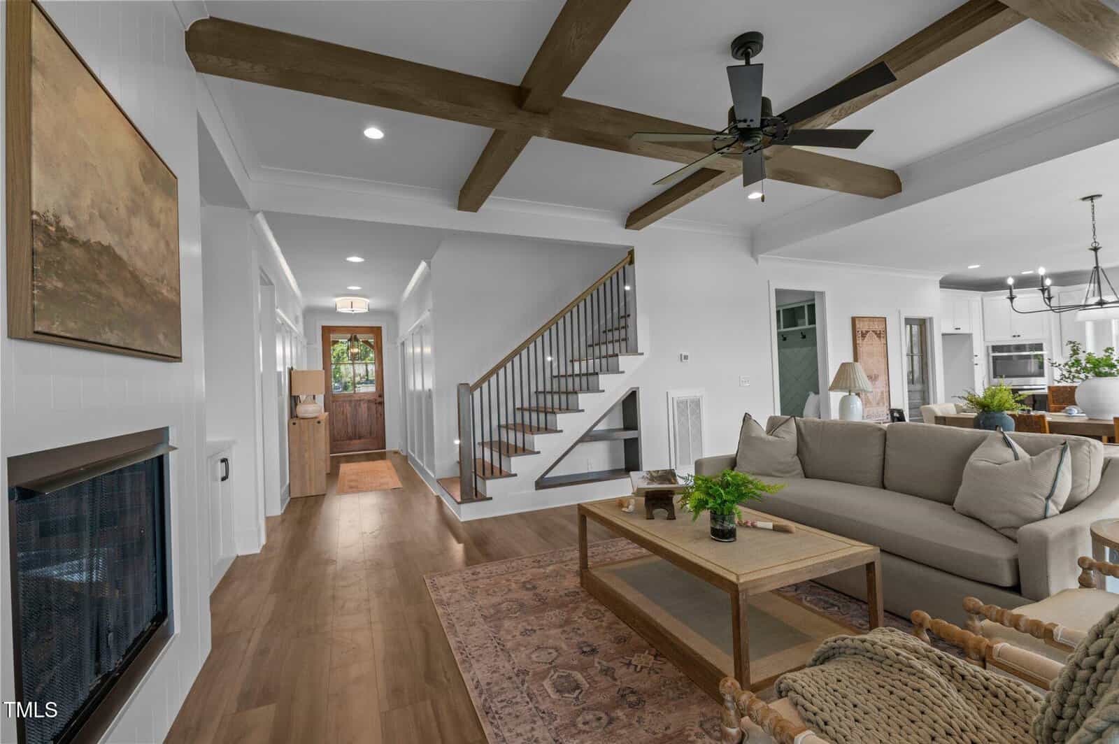 Living room with a beige couch, wooden ceiling beams, a ceiling fan, and a coffee table. Stairs lead to the second floor, and a hallway extends toward a front door with glass panels. Open kitchen and dining area in the background.