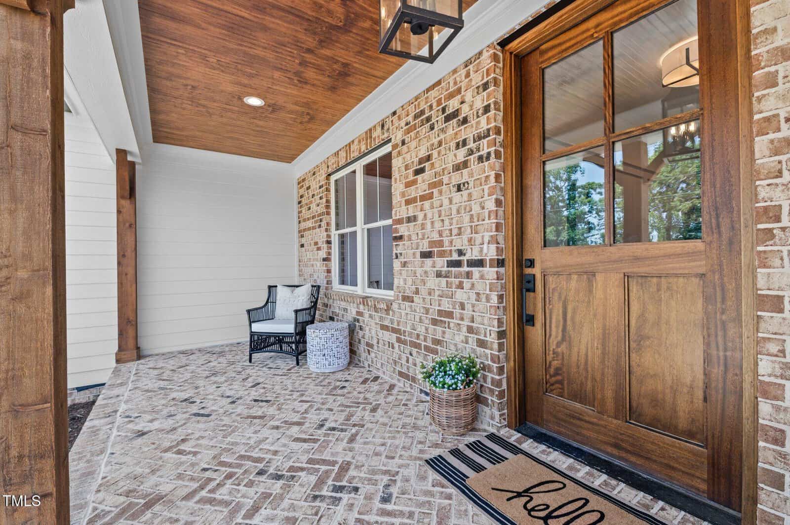 A brick-front porch with a wooden door, “hello” doormat, a potted plant, a black-and-white chair with cushions, and a wicker basket, all under a wooden ceiling.