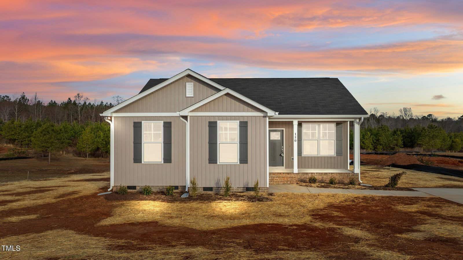 A single-story beige house with white trim and dark shutters sits on a dirt and straw yard, with a covered front porch and a walkway, against a backdrop of trees and a colorful sunset sky.
