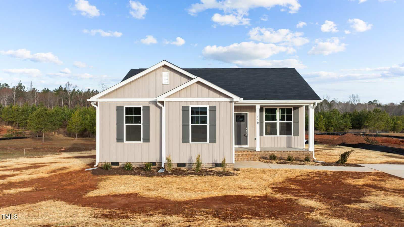 A small, single-story beige house with a black roof and white trim sits on a lot with fresh straw covering the ground. There are a few small plants by the front steps and trees in the background under a partly cloudy sky.