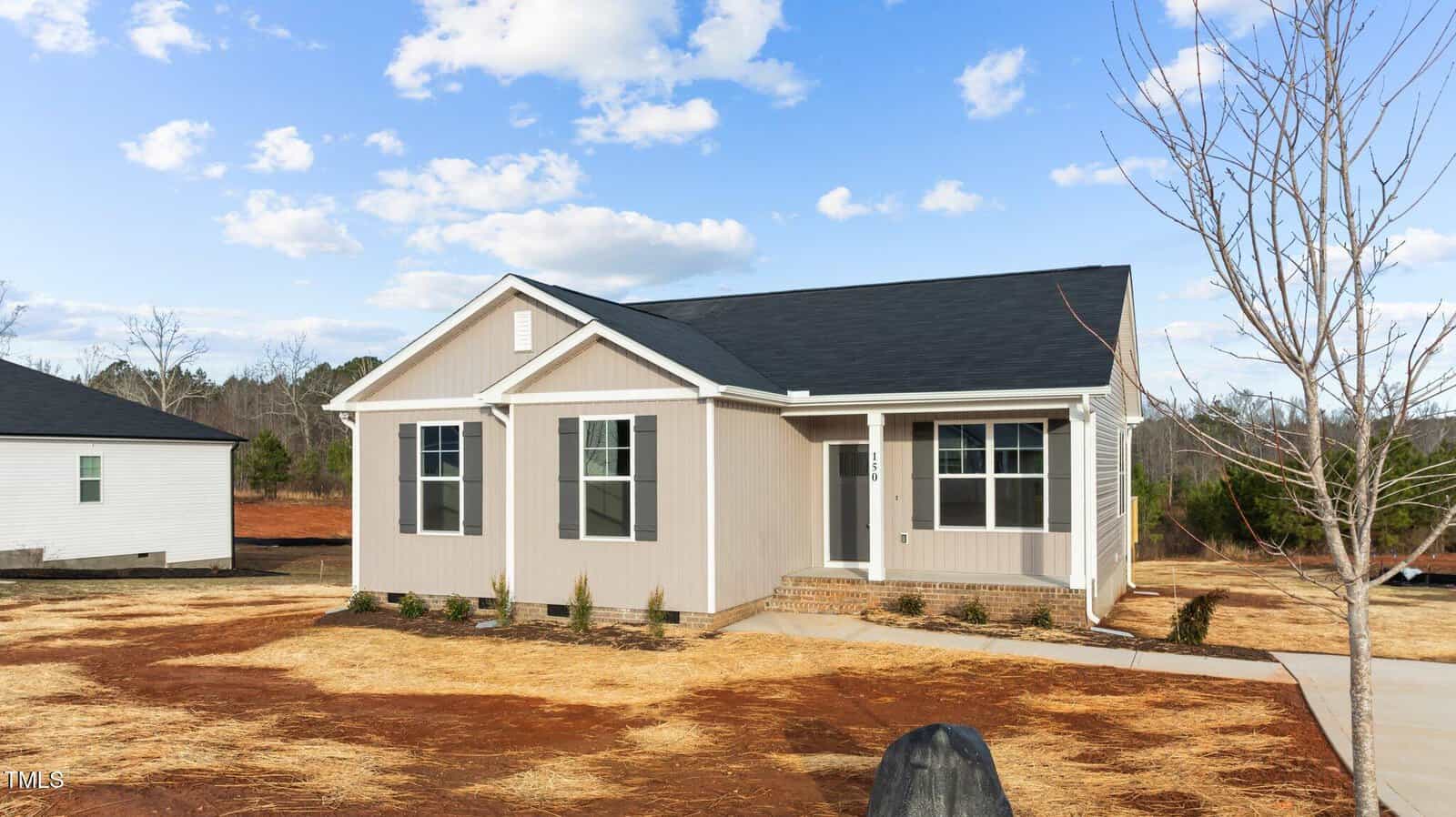A single-story beige house with white trim, black roof, and three front windows sits on a lot with straw-covered ground and minimal landscaping under a partly cloudy sky.