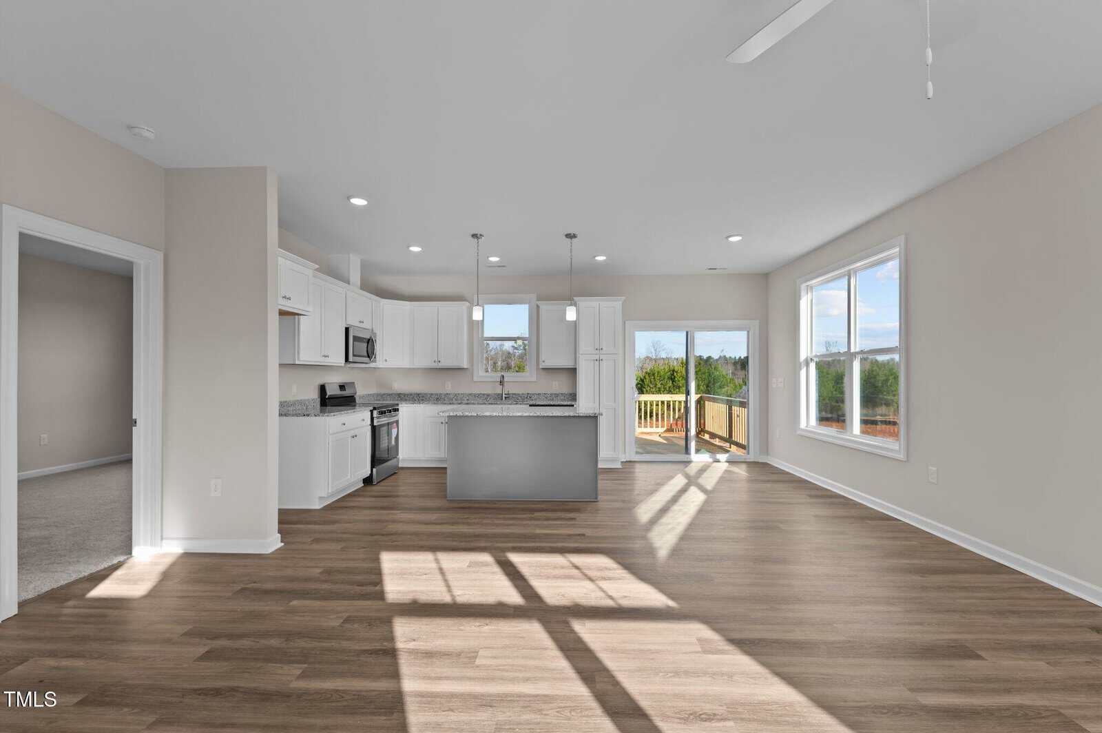 A modern, unfurnished open-concept kitchen and living area with white cabinets, stainless steel appliances, wood flooring, large windows, and sliding glass doors leading to a deck with a view of greenery outside.