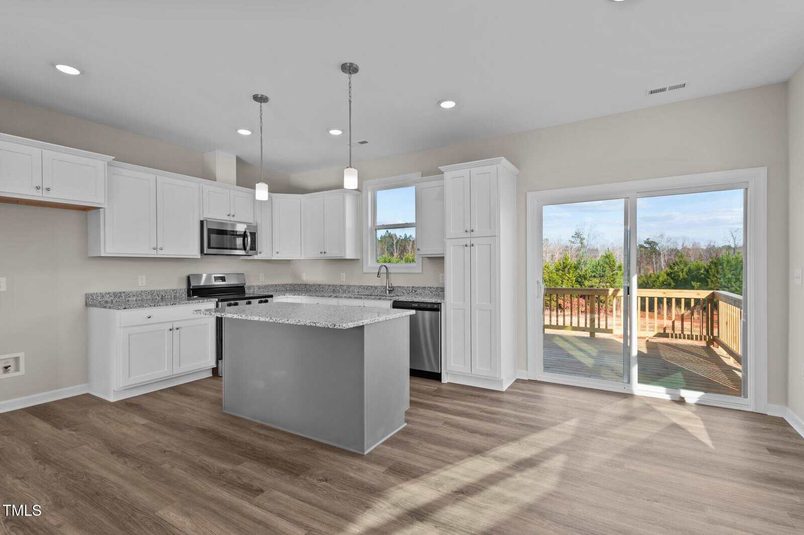 Modern kitchen with white cabinets, stainless steel appliances, a gray island with a granite countertop, light wood floors, pendant lights, and sliding glass doors leading to a wooden deck with a view of trees.