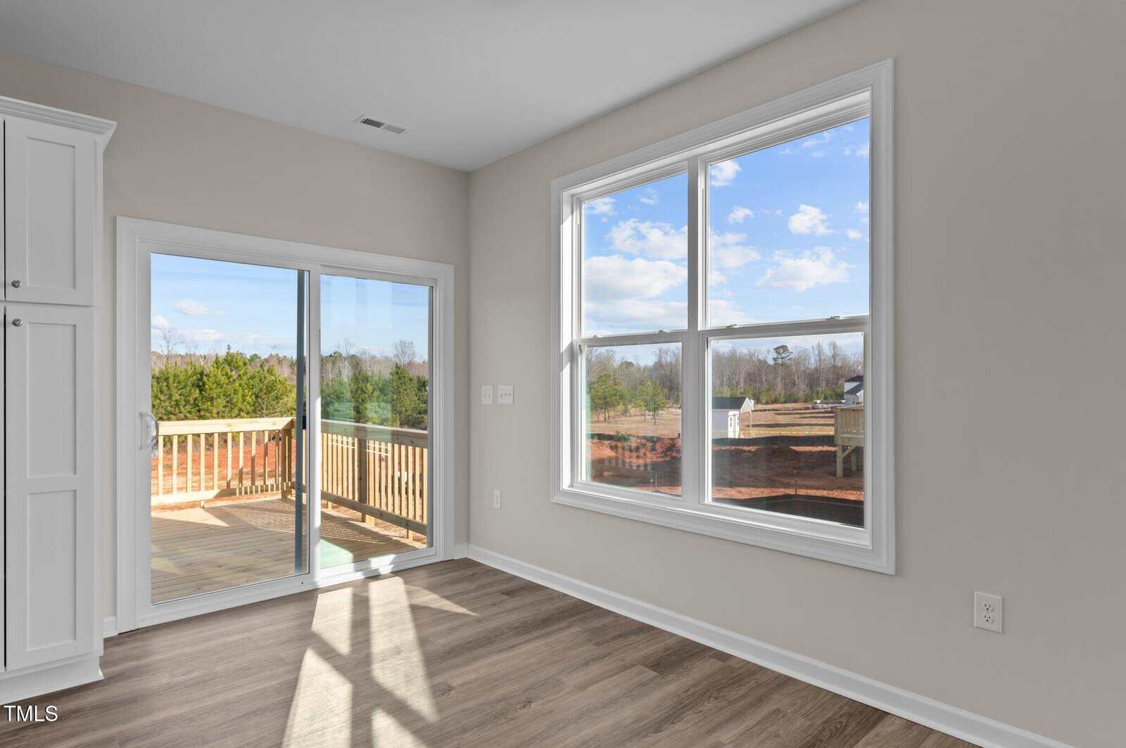 A bright room with light wood flooring, large windows, and a sliding glass door leading to a wooden deck. The view includes trees, a blue sky with clouds, and part of a red brick wall outside.