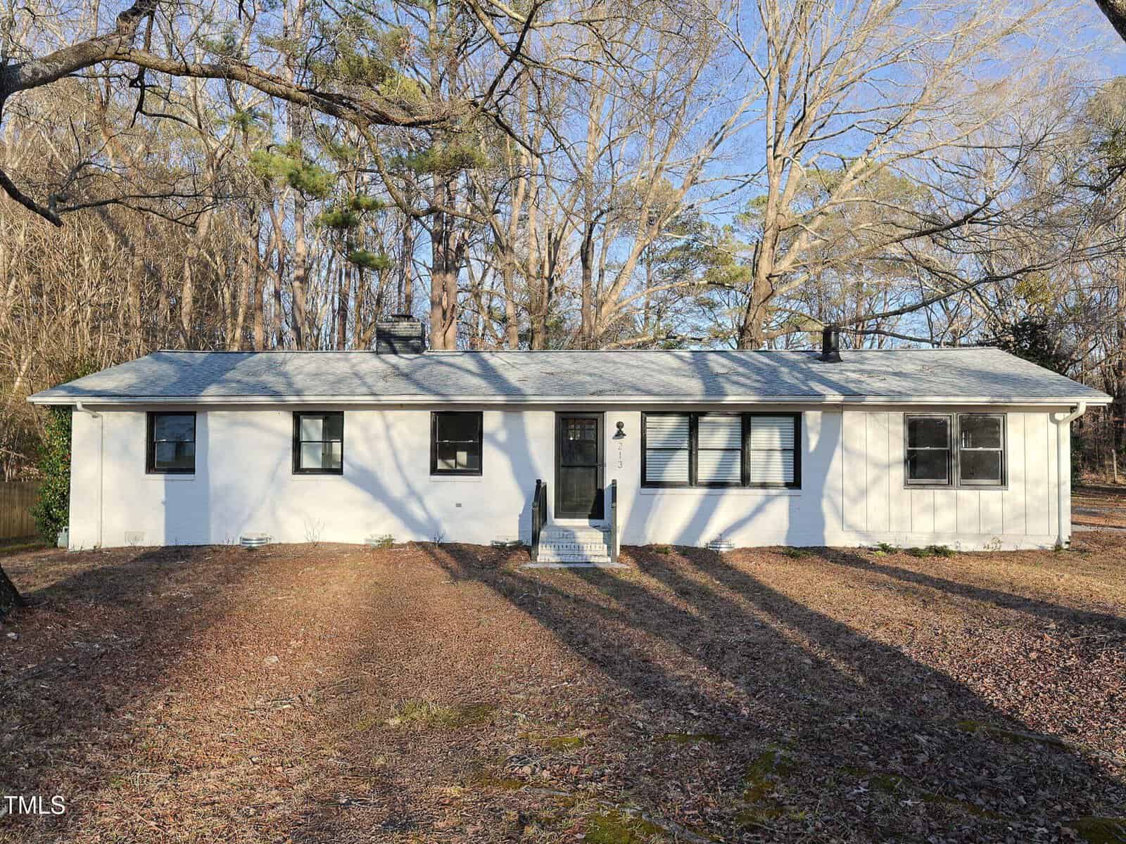 A single-story white house with a gray roof and black shutters, surrounded by leafless trees, sits on a lawn covered with dry leaves and dappled sunlight. There are steps leading up to the front door.