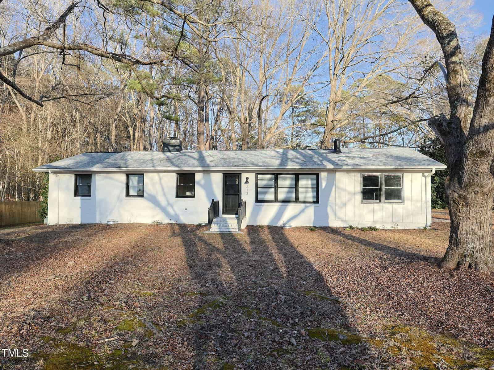 Single-story white house with a gray roof, black front door, and several windows. Leafless trees surround the yard, and shadows are cast across the ground on a clear day.