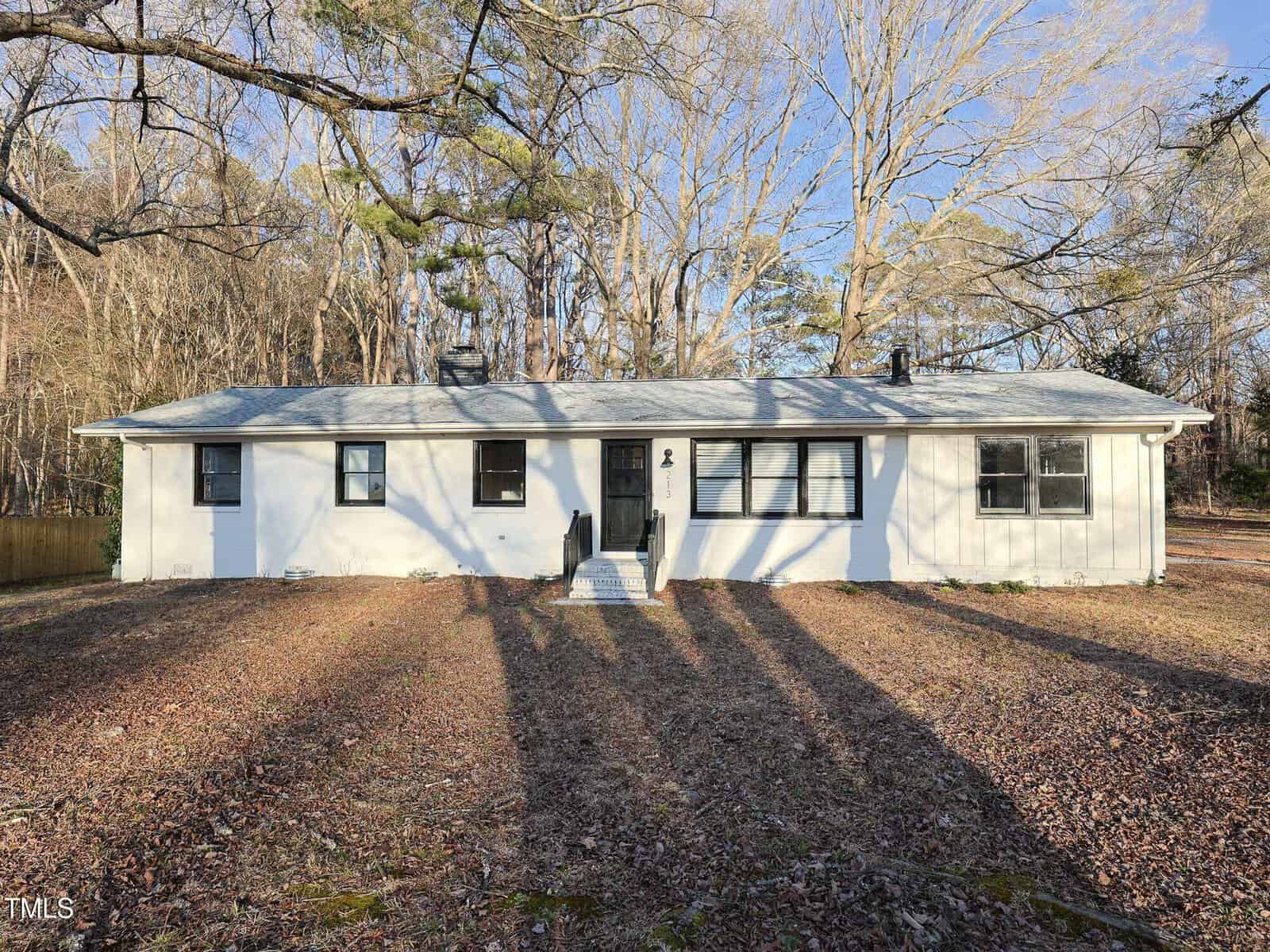 A single-story white house with a gray roof, black window frames, and a central front door. The yard is covered with dry leaves and bare trees, and long tree shadows stretch across the ground.