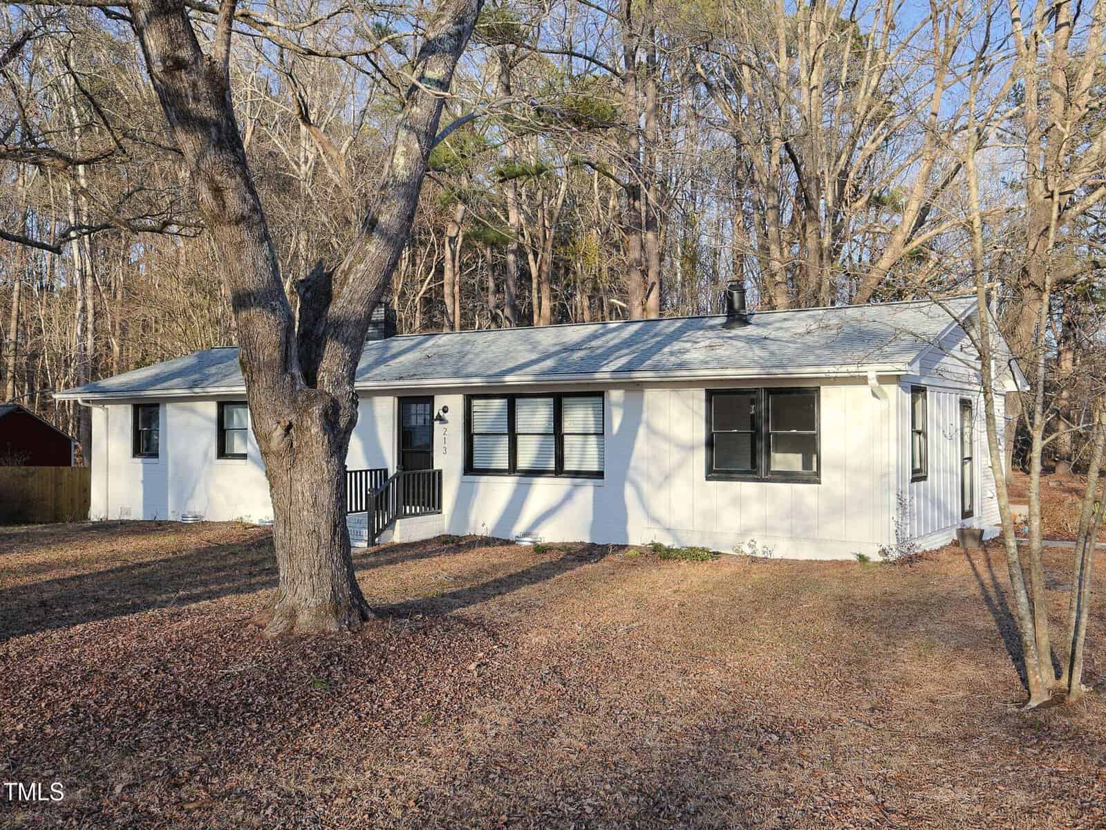 A single-story white house with black trim and a small front porch, situated on a lawn covered with fallen leaves and surrounded by leafless trees. The background features dense woods.
