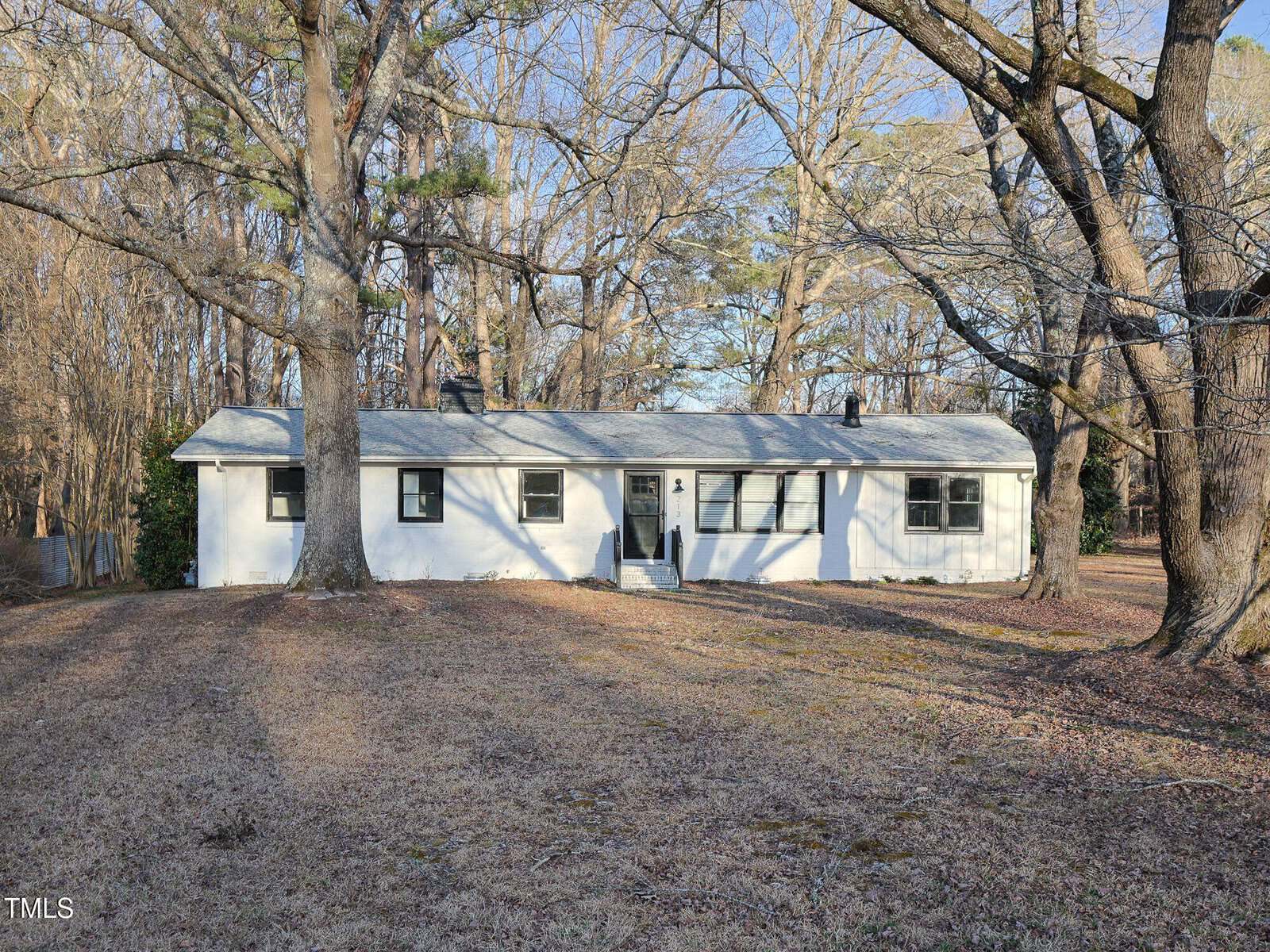 Single-story white house with a gray roof, black shutters, and a small front stoop. Large trees with bare branches surround the house, and the yard is covered in brown grass. The scene appears to be in winter or early spring.