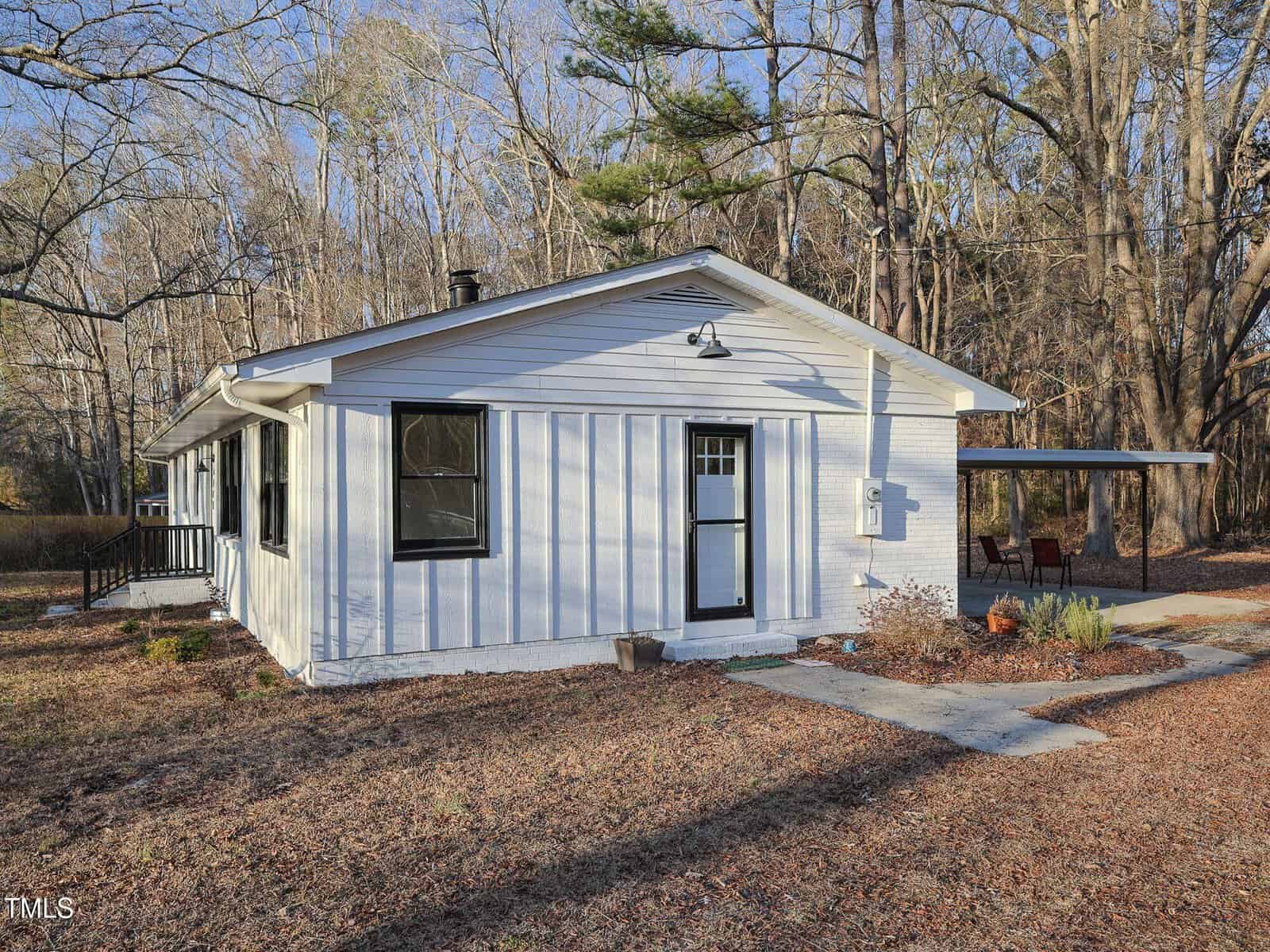 Single-story white house with black trim, a front door with a glass panel, and adjacent covered carport. The house sits on a lot with leaf-covered ground and is surrounded by leafless trees. A concrete walkway leads to the entrance.