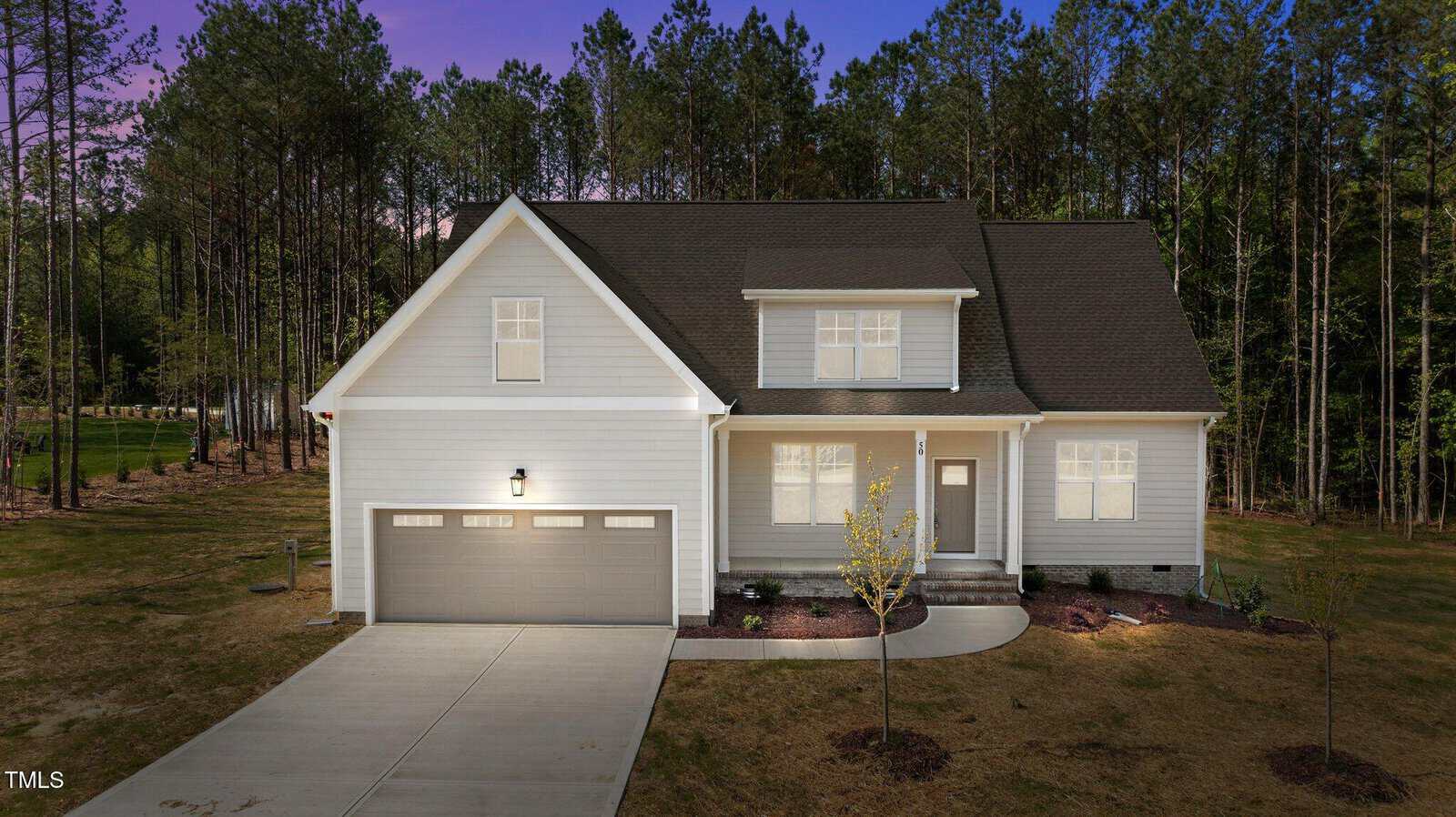 A two-story, light gray house with a dark roof, double garage, and front porch is set on a large lot with minimal landscaping, surrounded by tall pine trees at sunset.
