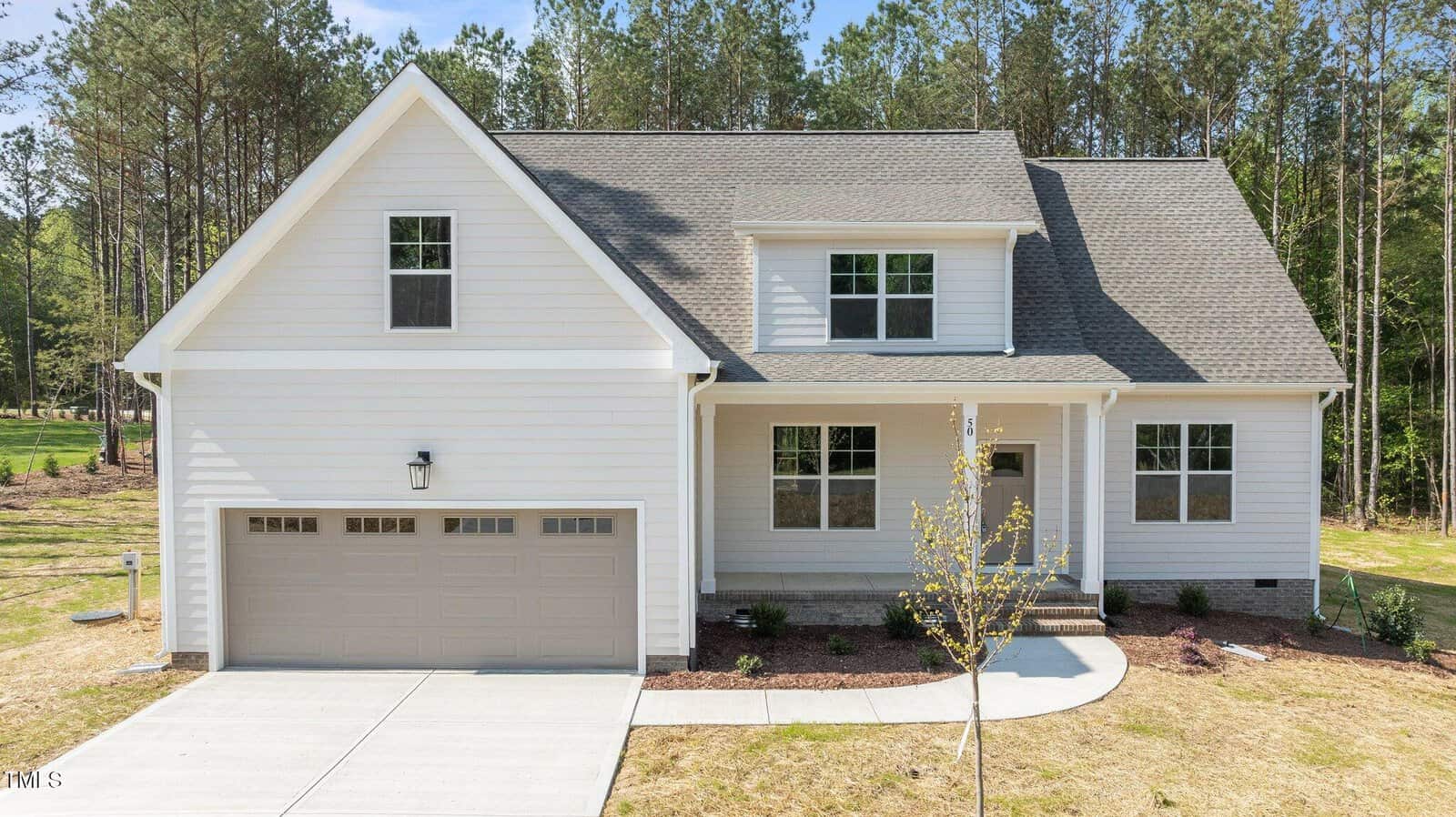 A two-story house with light-colored siding, a double garage, and a covered front porch. The yard has young landscaping, and the home is surrounded by tall trees in the background.