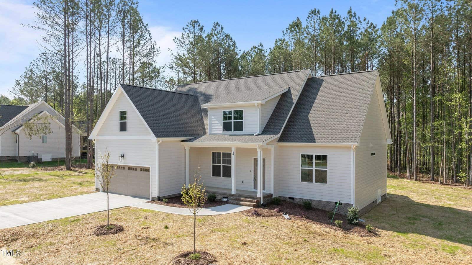 A modern two-story house with white siding, a covered front porch, attached two-car garage, and new landscaping, set on a grassy lot with pine trees in the background under a clear sky.