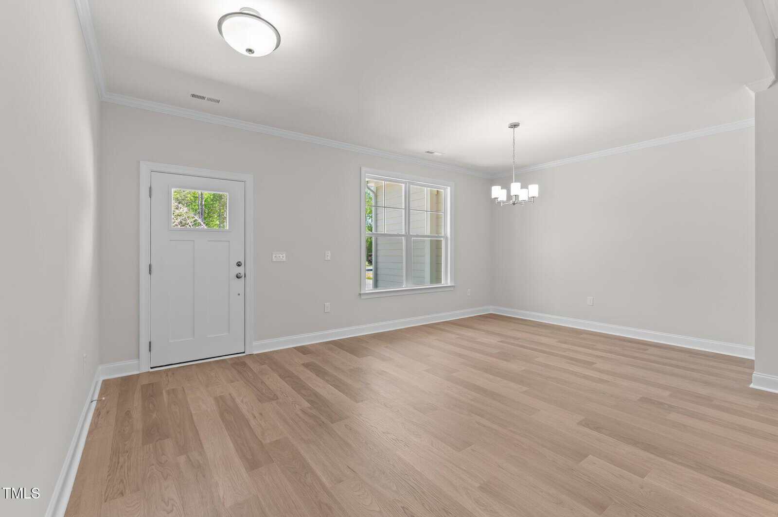 Empty room with light wood floors, light gray walls, white trim, a white front door, a large window, a ceiling light near the door, and a modern chandelier hanging from the ceiling.