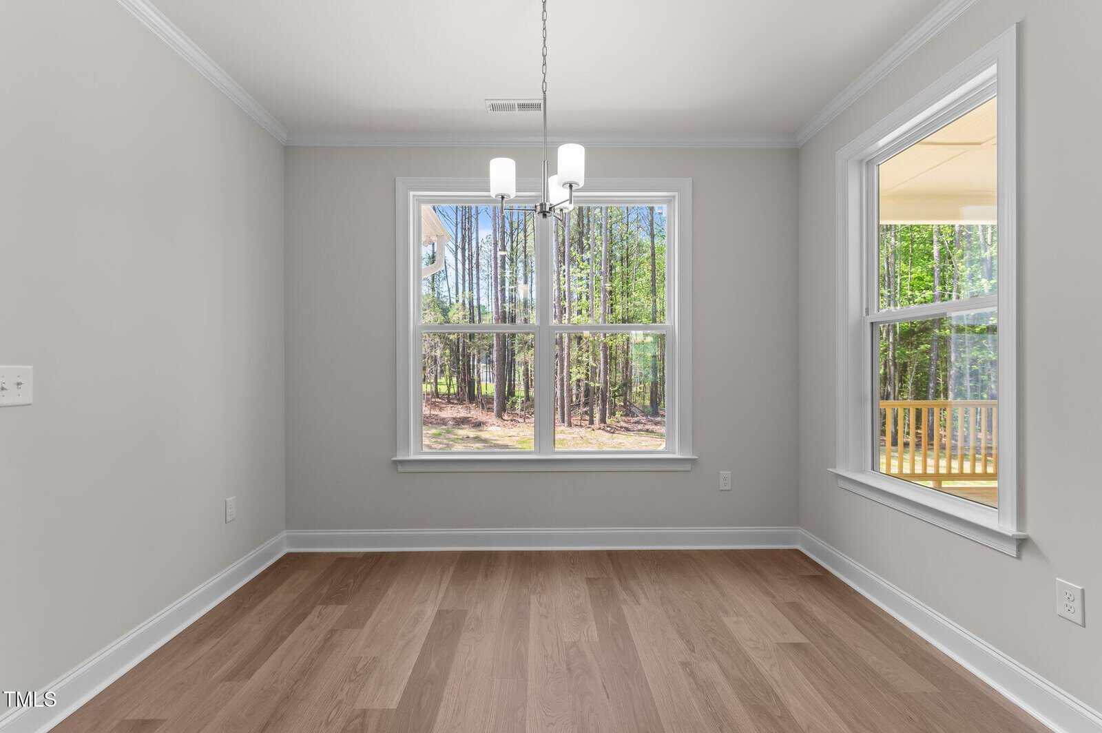 Bright dining area with light wood flooring, light gray walls, a white ceiling, two large windows overlooking trees, and a modern ceiling light fixture. A porch is visible through the right window.