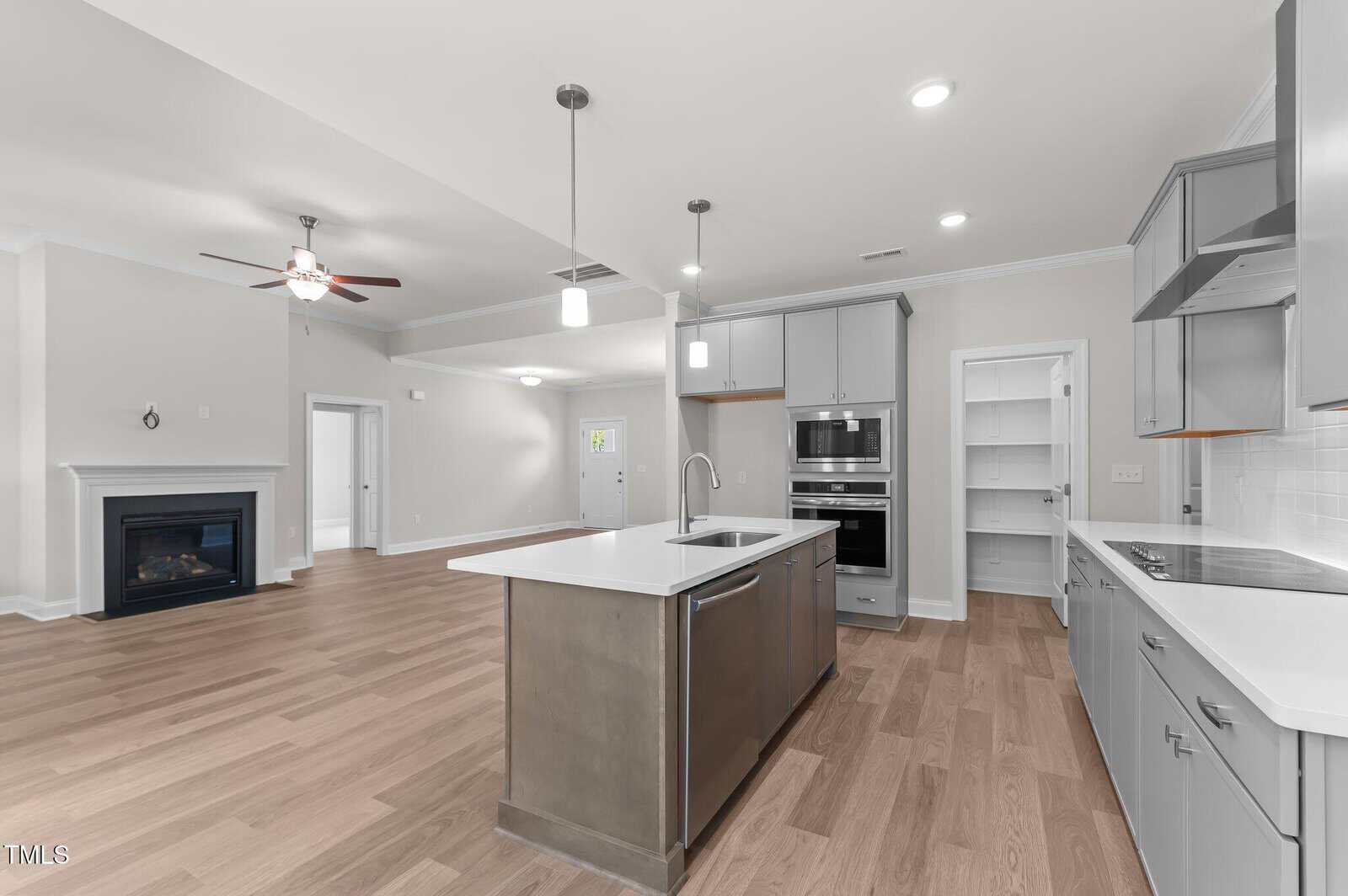 Modern kitchen with gray cabinets, a center island with sink, built-in oven and microwave, open shelving, and light wood flooring. The space opens to a living area with a ceiling fan and a fireplace on the left.