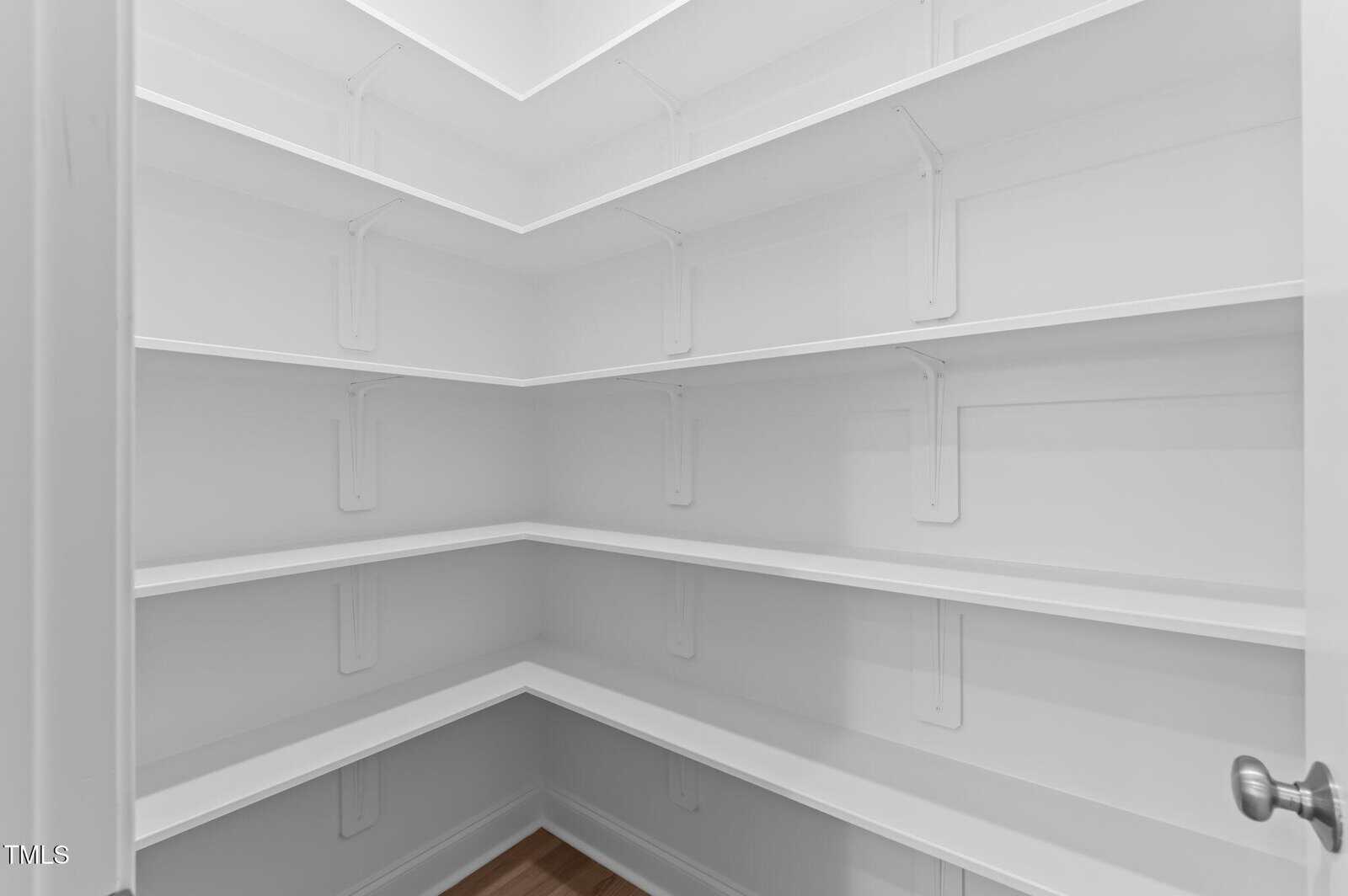 Empty white walk-in pantry with four rows of shelves along two walls, white brackets supporting the shelves, and a wooden floor. The photo is taken from the entrance of the pantry, showing the shelving layout.