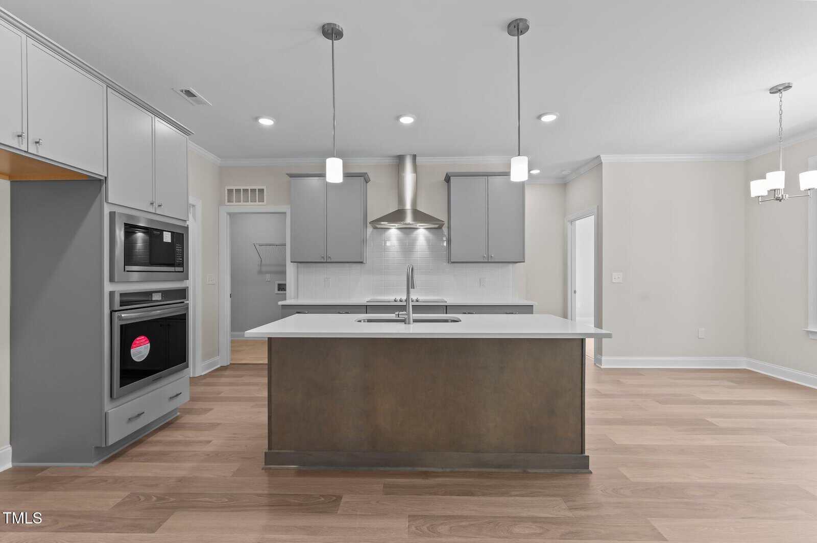 A modern kitchen with light wood floors, a central island with a sink, light gray cabinets, stainless steel appliances, pendant lights above the island, and a chandelier in the adjacent dining area.