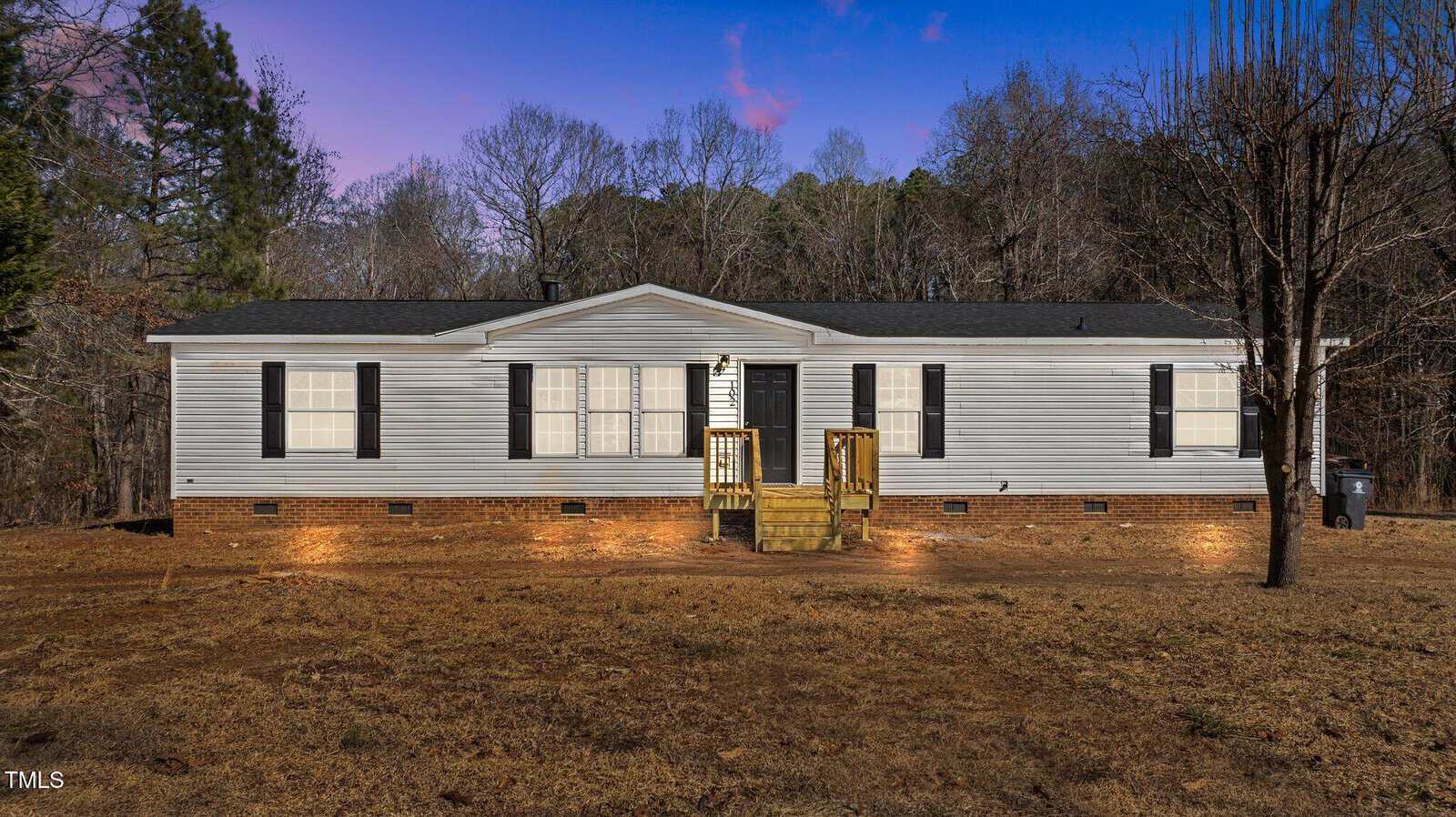 A single-story manufactured home with white siding, black shutters, and a small wooden porch sits on a dry, brown lawn, with leafless trees and forest in the background at dusk. Outdoor lights illuminate the house.