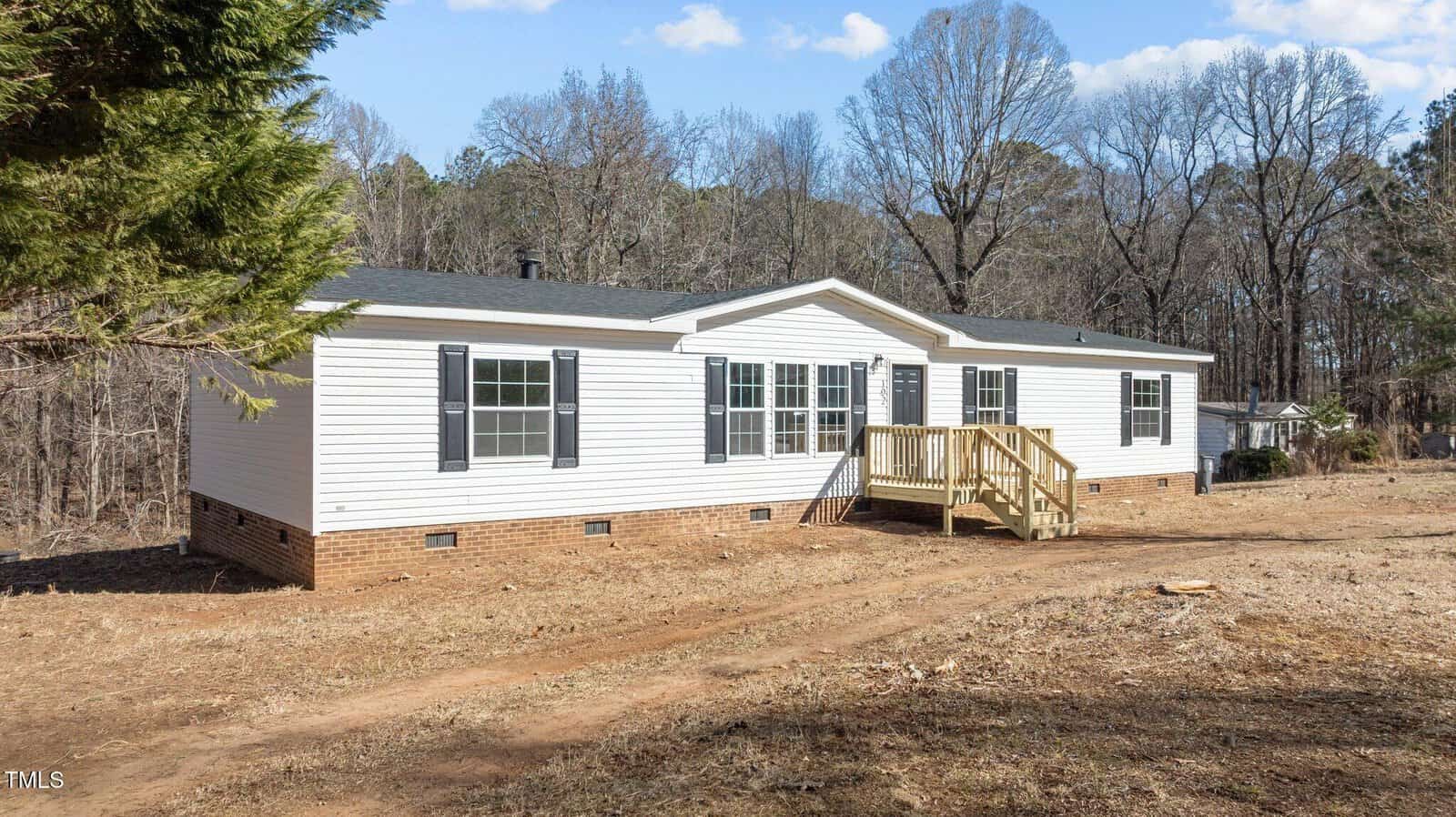 A white, single-story manufactured home with black shutters and a small wooden porch sits on a dirt lot, bordered by trees and sparse grass, under a partly cloudy sky.