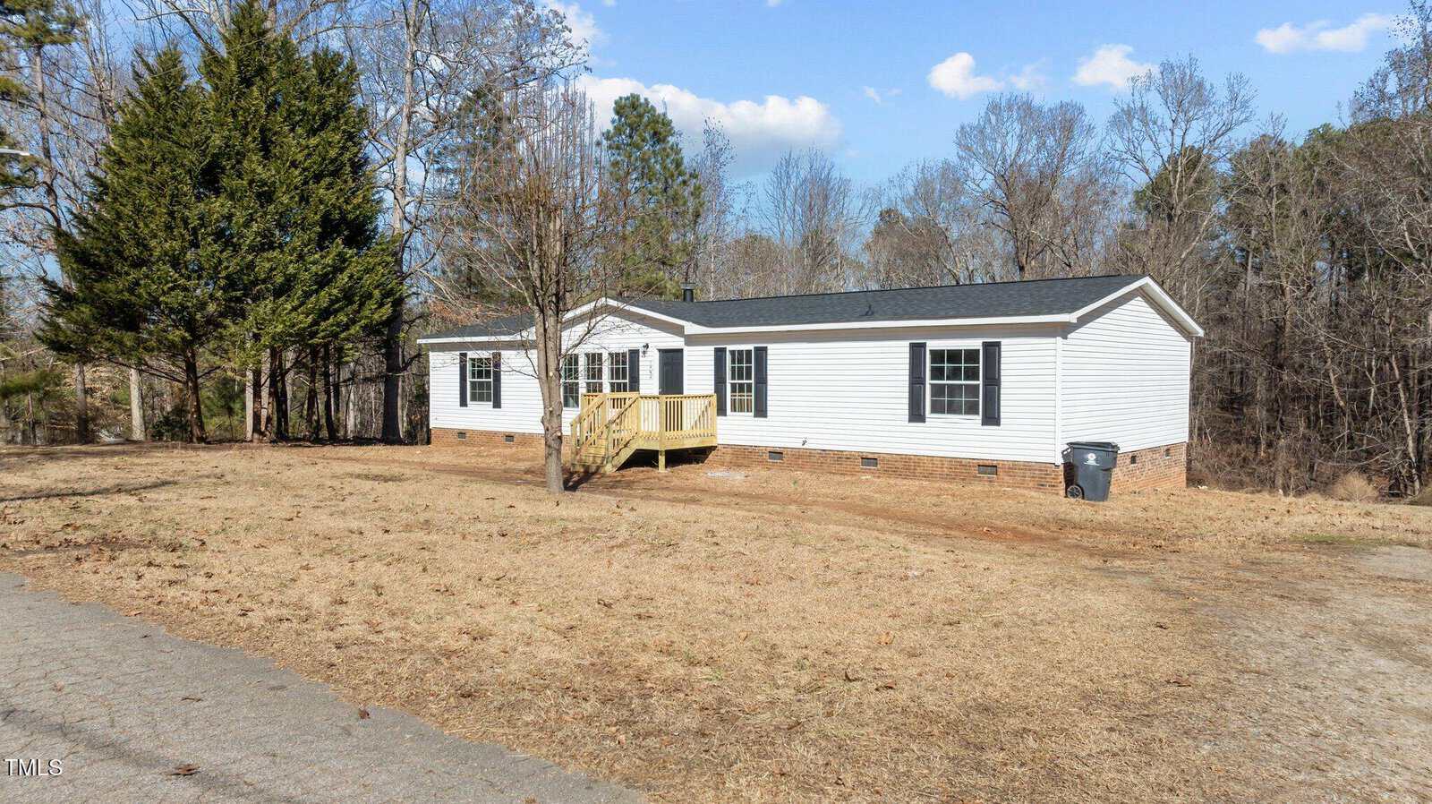 A single-story white manufactured home with black shutters and a small wooden front porch sits on a dry, grassy lot with sparse trees and a paved driveway under a partly cloudy sky.