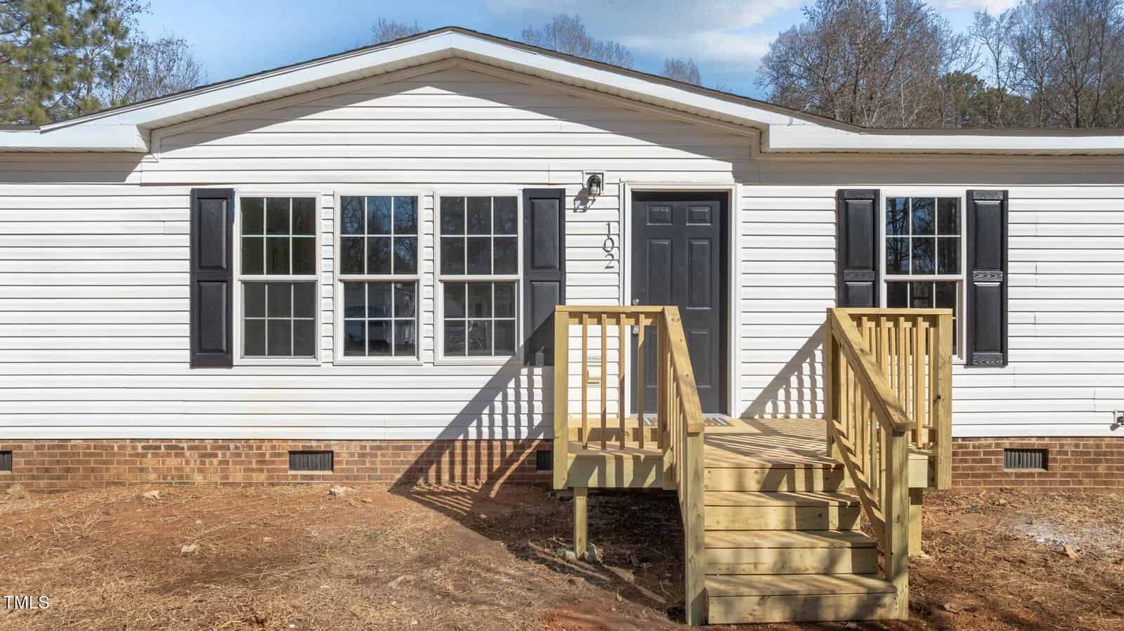 A white, single-story manufactured home with black shutters, a central dark front door, and a newly built wooden ramp and steps leading to the entrance. The yard is bare dirt, and trees are visible in the background.