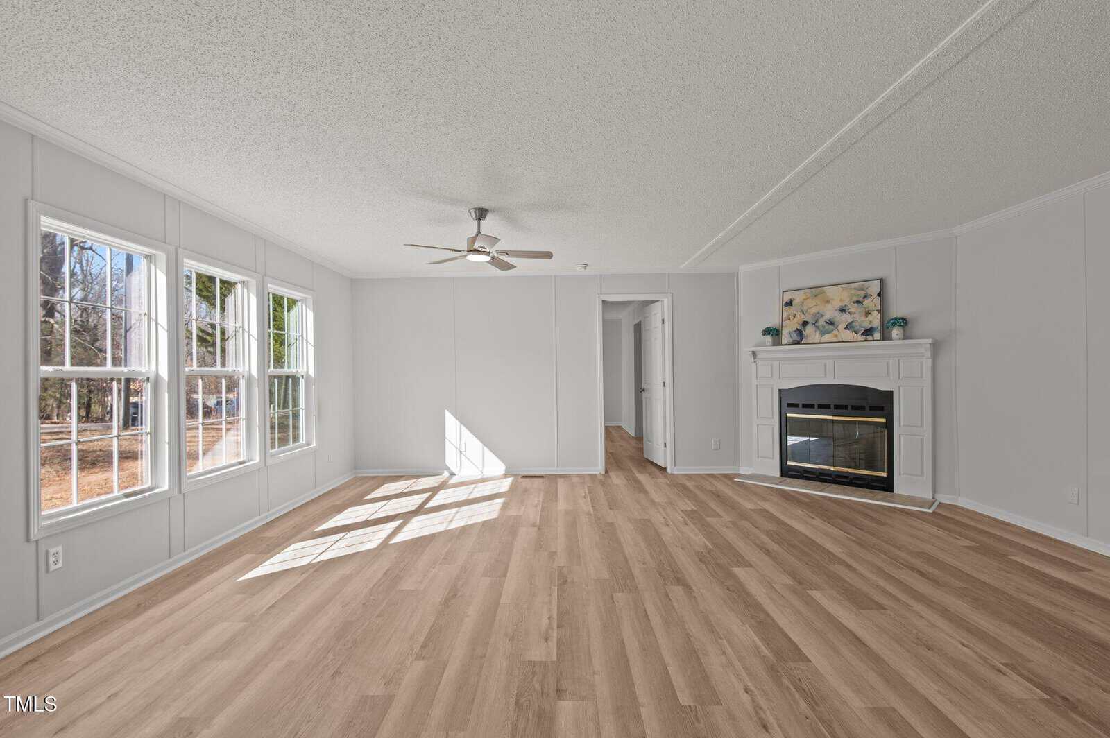 A bright, empty living room with light wood floors, white walls, three large windows, a ceiling fan, and a white fireplace with artwork above it. Sunlight streams in through the windows onto the floor.