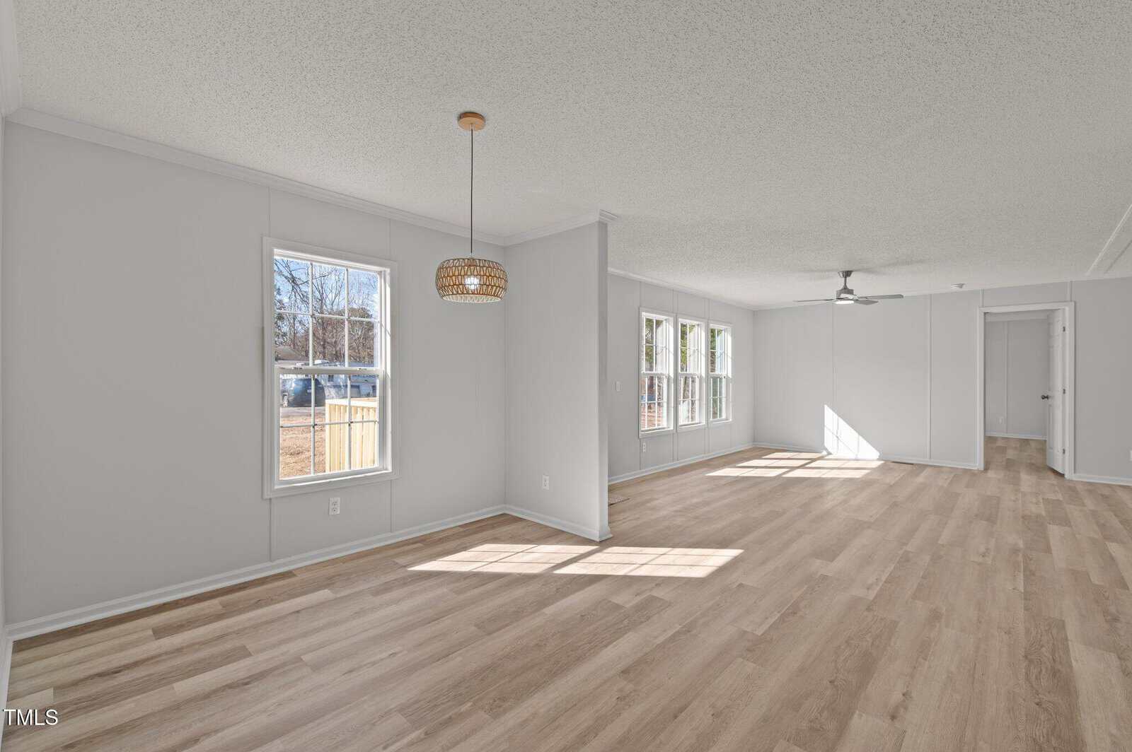 A spacious, empty living area with light wood flooring, white walls, large windows, a ceiling fan, and a pendant light fixture. Sunlight enters the room, and a hallway leads to other parts of the house.