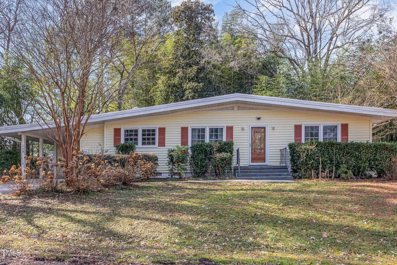 A single-story house with light yellow siding, red shutters, a covered carport on the left, front steps leading to a wooden door, and mature trees and shrubs in the front yard.