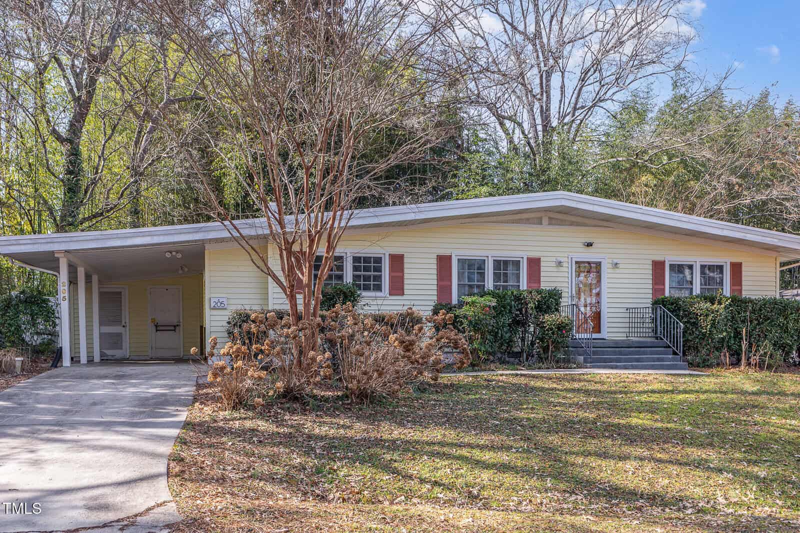 Single-story yellow house with red shutters, a covered carport on the left, a front porch with black railings, and a concrete driveway. Leafless trees and shrubs are in the yard, and tall trees surround the house.