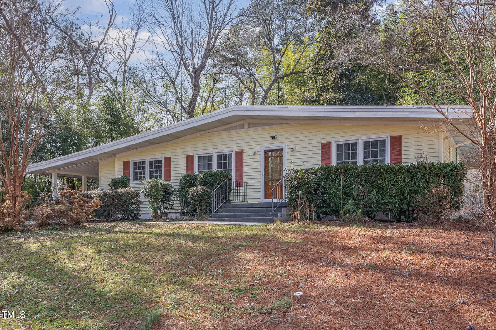 Single-story house with pale yellow siding, red shutters, and a dark front door, set behind shrubs and trees. The yard features patchy grass and pine straw, with bare trees and greenery in the background.