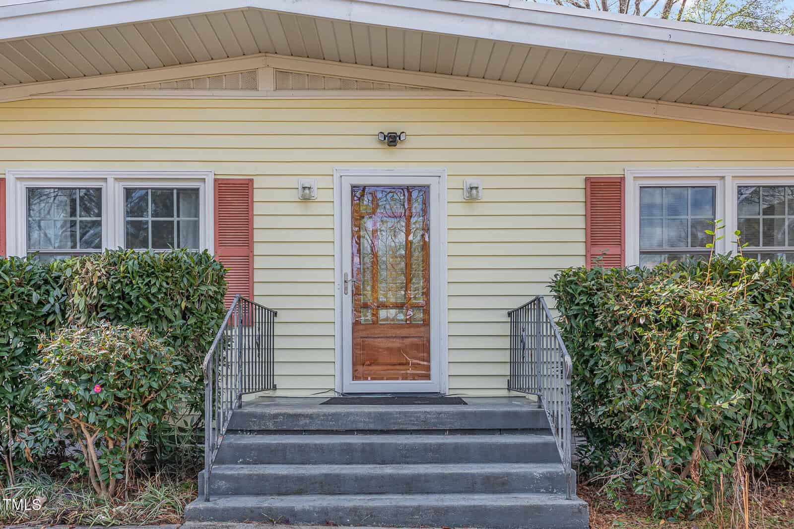 A front entrance of a yellow house with white trim, two windows with reddish shutters on either side of a glass door, metal railings on both sides of concrete steps, and bushes flanking the walkway.