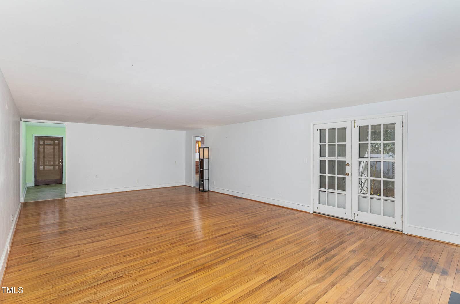 Spacious empty room with light-colored walls, hardwood floors, and French doors leading outside. A tall lamp is placed near the doors, and there is a wooden door visible at the far end of the room.