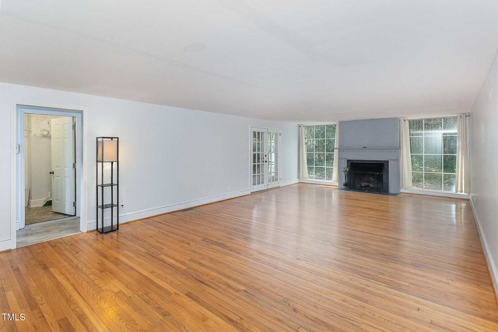 Spacious empty living room with hardwood floors, a gray fireplace, large windows, a floor lamp, and a doorway leading to a bathroom. Walls are painted white, and natural light fills the room.