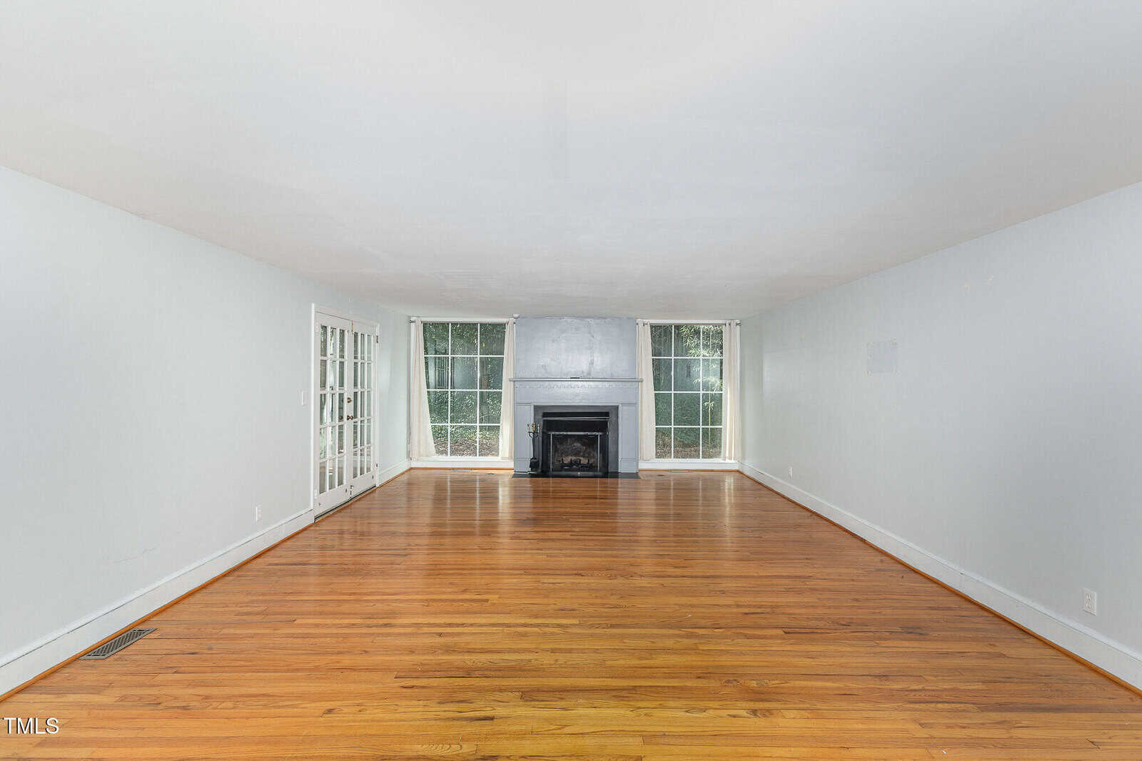 Empty living room with hardwood floors, white walls, a fireplace centered on the far wall, large windows flanking the fireplace, and a set of glass French doors on the left side.