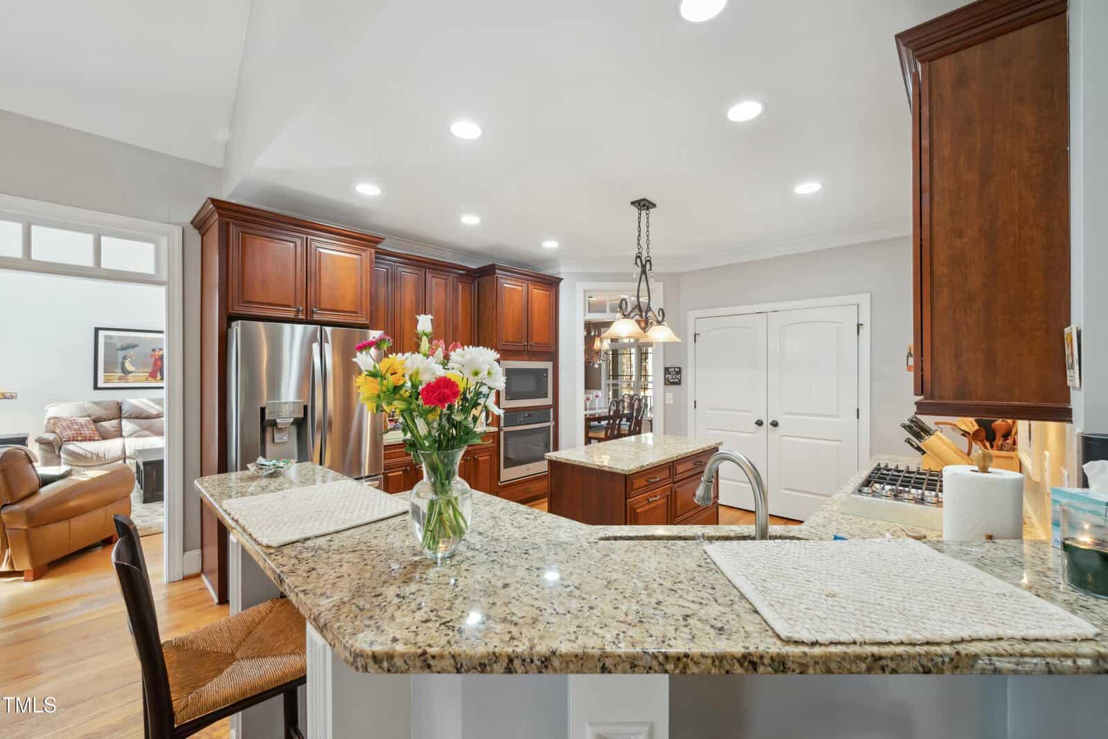 Modern kitchen with granite countertops, wooden cabinets, stainless steel appliances, and a center island. A vase with colorful flowers sits on the counter. Recessed ceiling lights brighten the space. Living room visible in background.