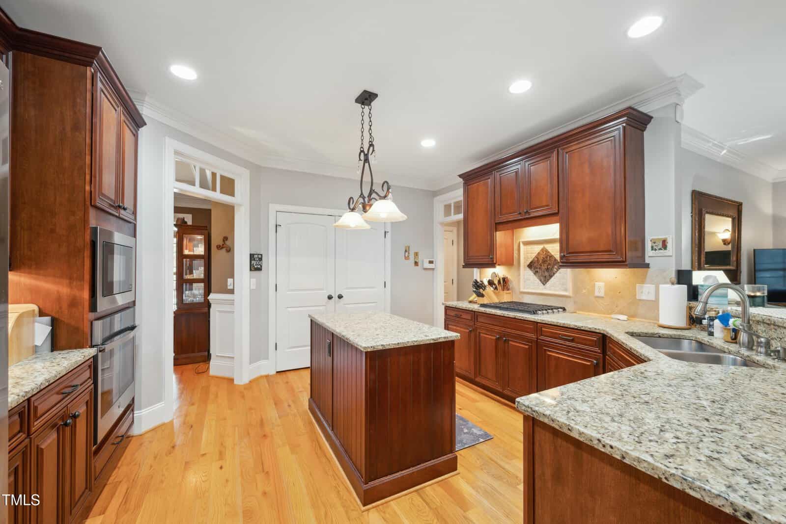 Spacious kitchen with wood cabinets, granite countertops, an island, stainless steel appliances, light hardwood floors, and hanging pendant lights. Doors and archways lead to other rooms in the house.
