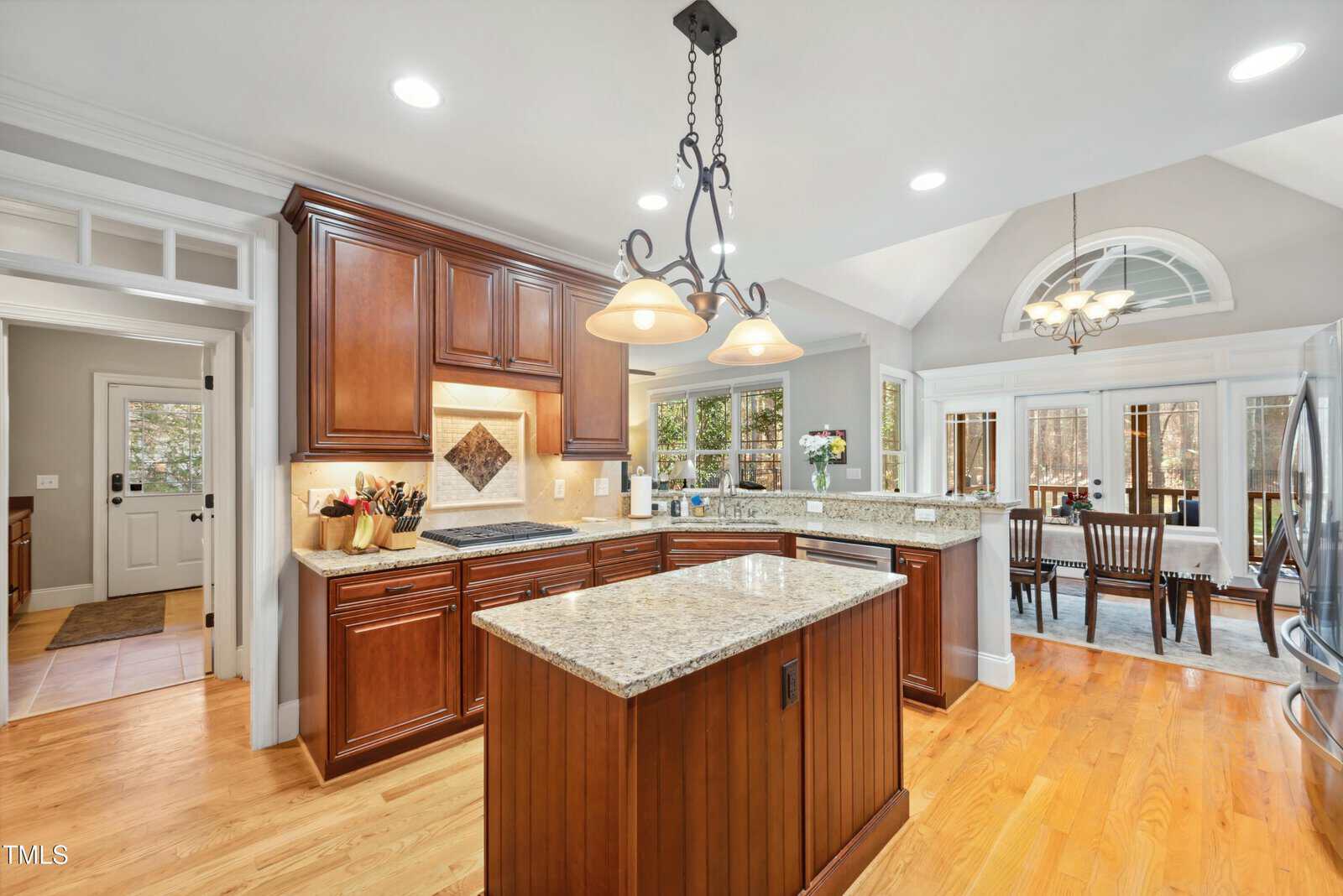 A kitchen with wooden cabinets, granite countertops, a center island, stainless steel appliances, and pendant lighting. An open dining area with large windows and a vaulted ceiling is visible in the background.