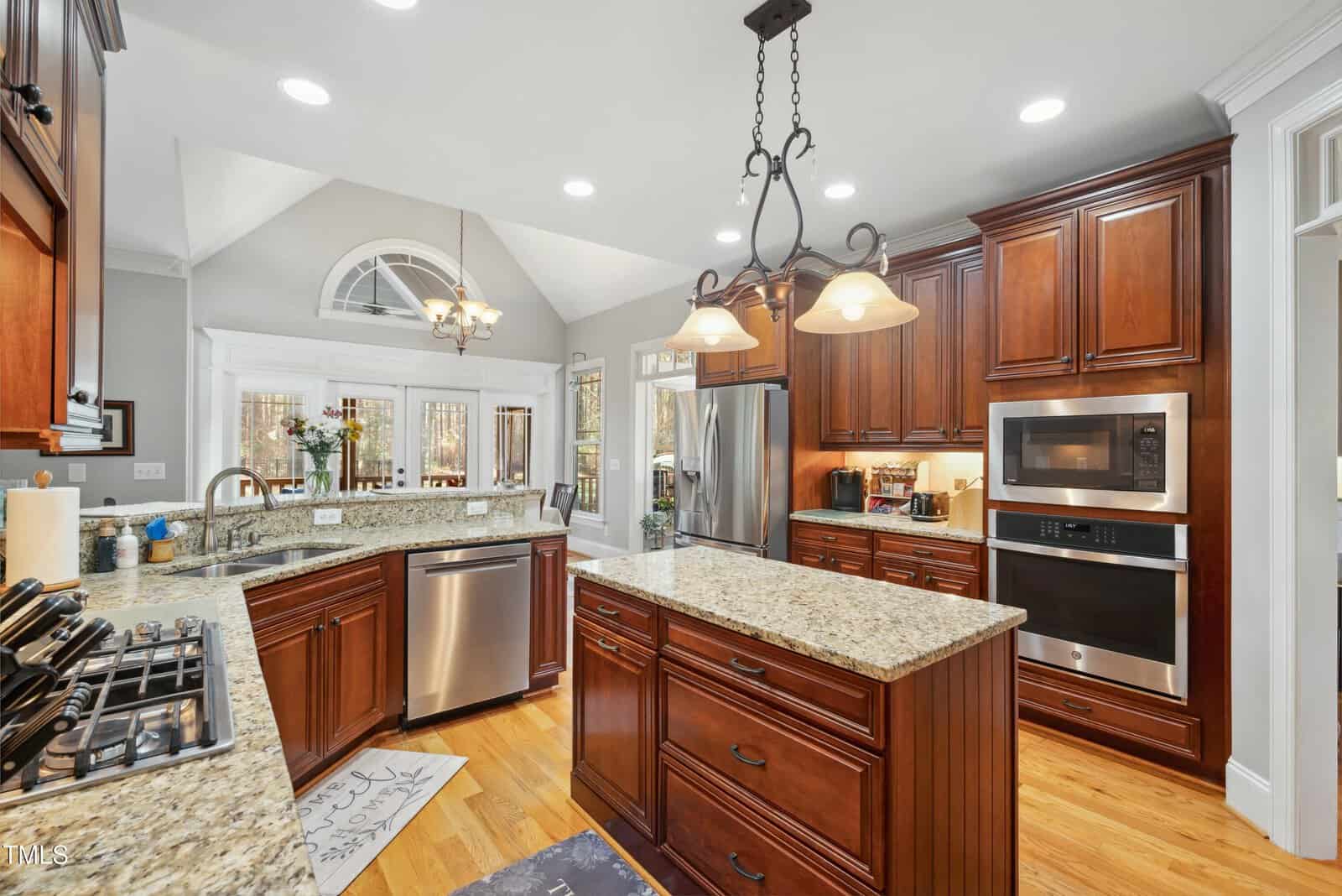 Spacious kitchen with wood cabinets, granite countertops, stainless steel appliances, a central island, double ovens, hanging lights, and hardwood floors. Large windows and skylights let in natural light.
